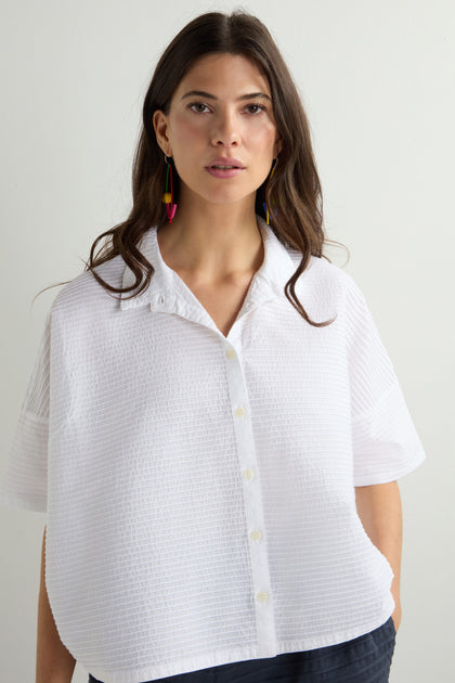 A woman with long brown hair models a Pleated Short Boxy Shirt made of luxurious cotton lawn, paired with colorful dangly earrings, set against a plain light background.