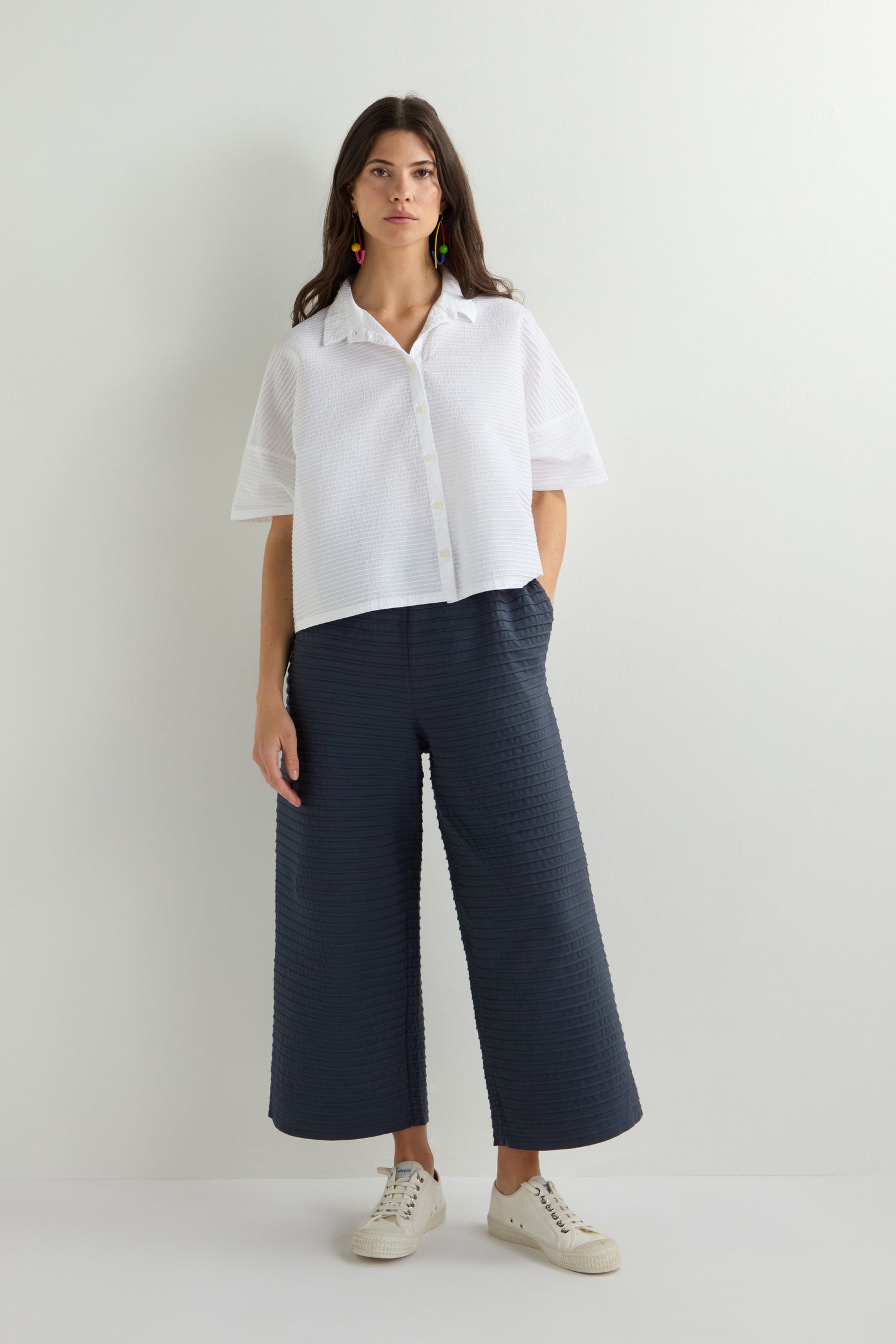 A woman in a Yacco Maricard Pleated Short Boxy Shirt with dark wide-leg pants and white sneakers stands against a plain background.