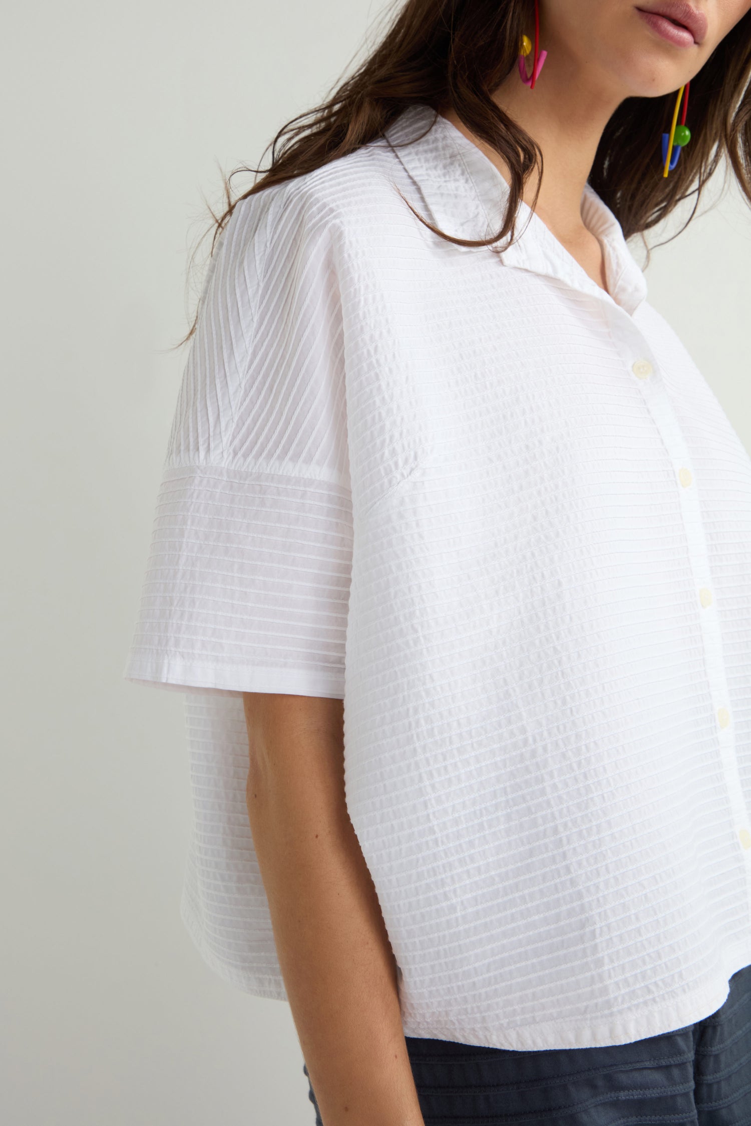 Against a plain background, someone wears Yacco Maricard's elegant Pleated Short Boxy Shirt in pristine white. Their vibrant earrings provide a lively touch, highlighting the brand's signature style.