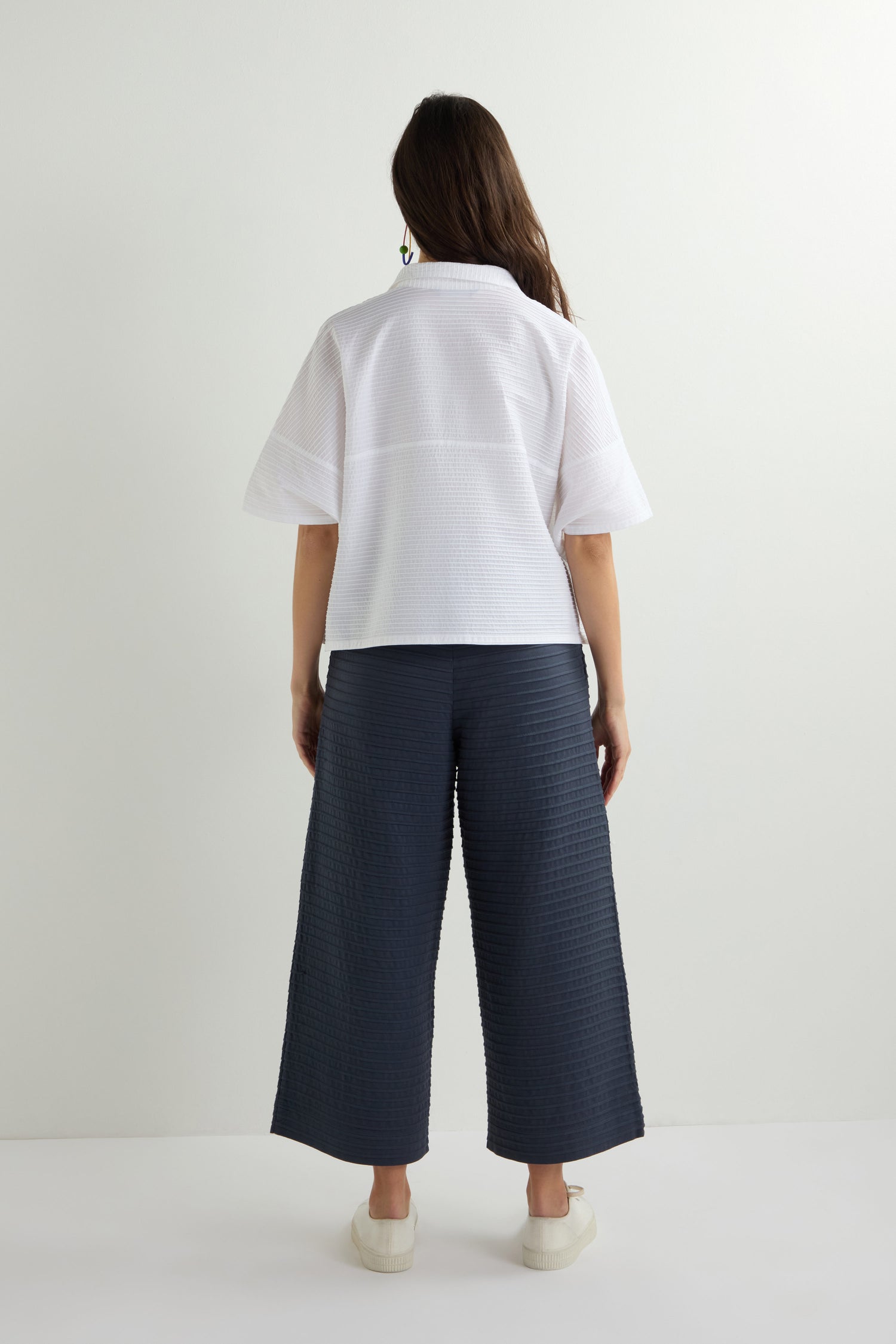 Wearing a Pleated Short Boxy Shirt, a person exudes understated elegance in this white short-sleeve top paired with dark wide-leg pants. The luxurious cotton lawn offers an airy feel, while pristine white sneakers stand out against the plain background.