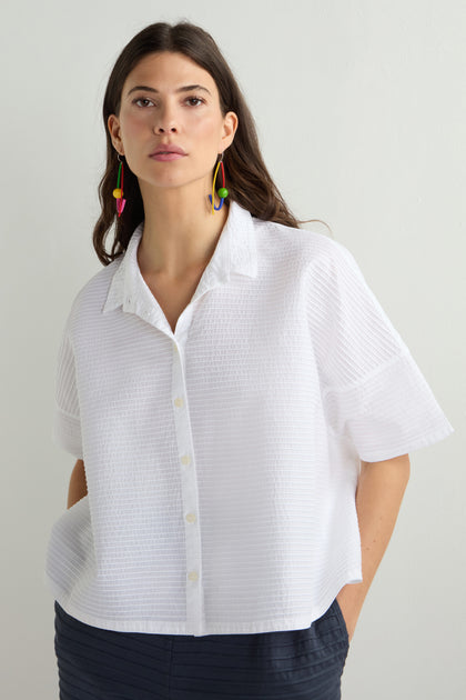 A person with long hair is wearing a luxurious Pleated Short Boxy Shirt and colorful earrings, standing against a plain background.