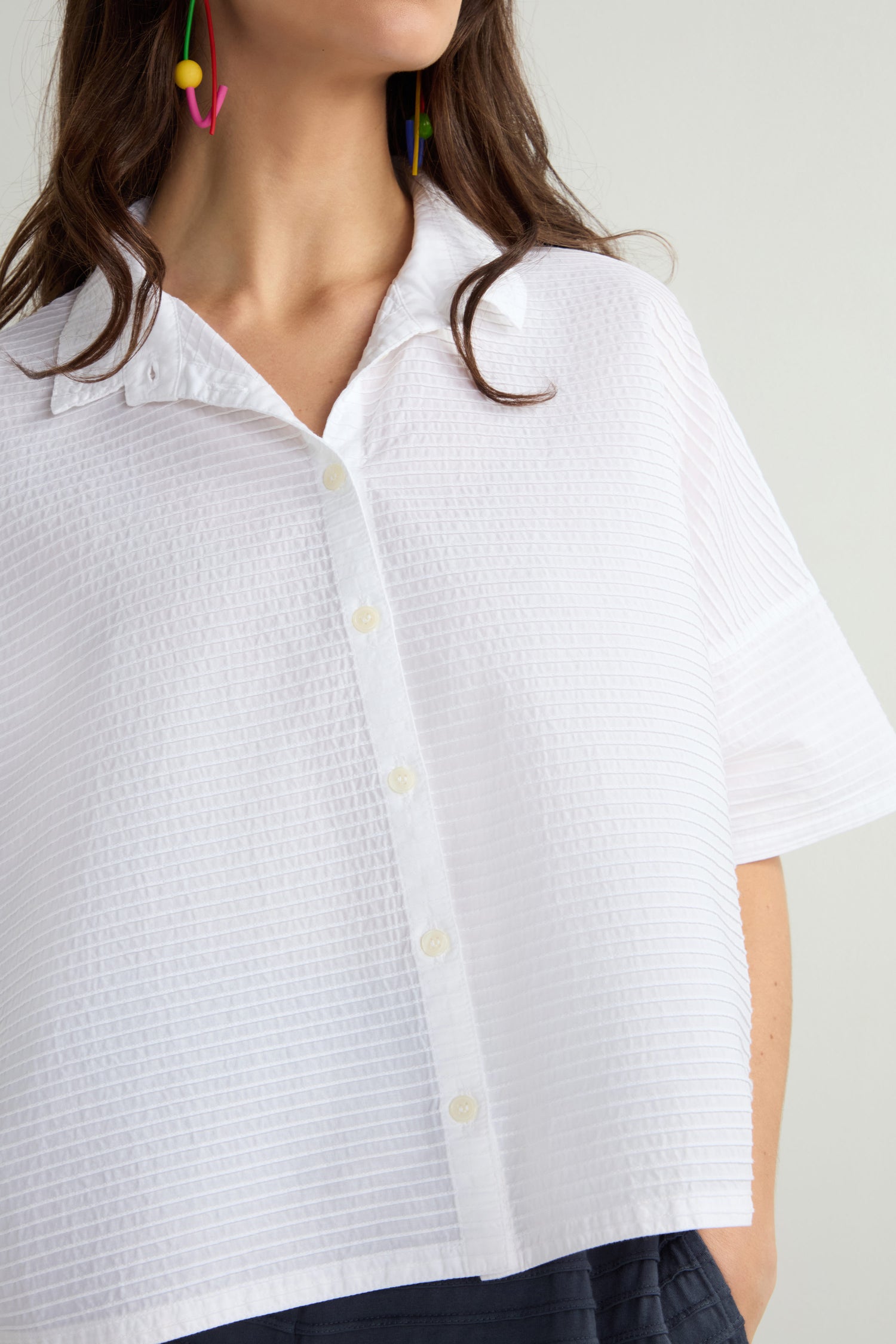 Someone is wearing a luxurious white short-sleeve button-up shirt, similar to Yacco Maricard's Pleated Short Boxy Shirt, featuring a subtle texture. Colorful earrings and dark pants complement the look.