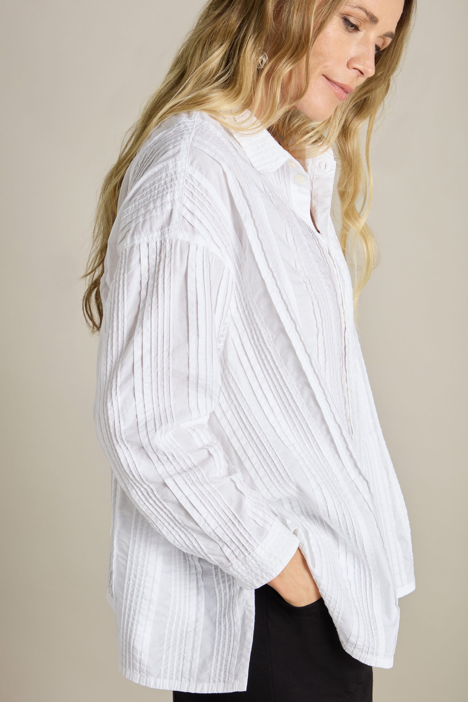 A person with long blonde hair wearing a white Multi Pleat Shirt from Yacco Maricard and black pants, looking down with hands in pockets against a plain background.