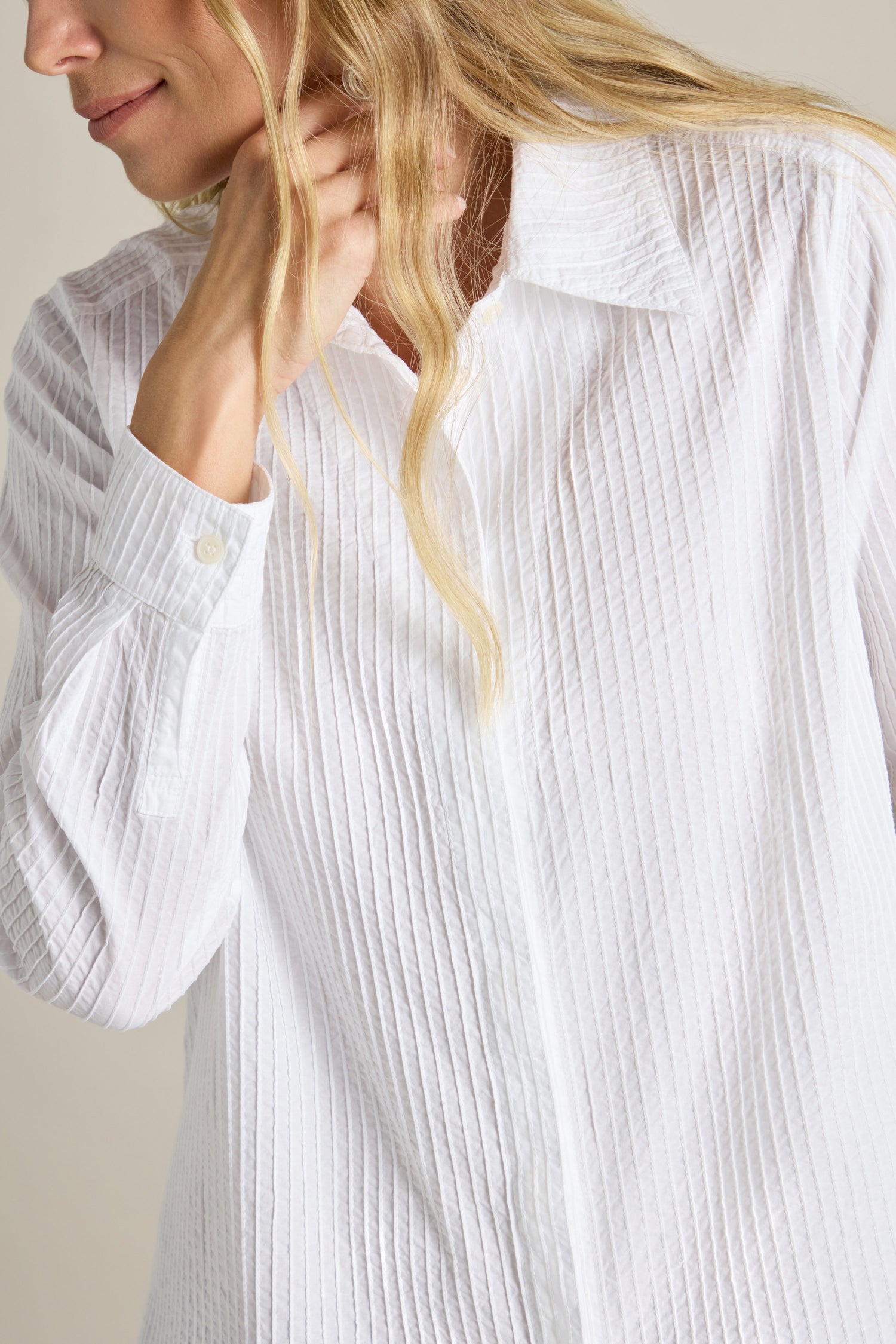 A person wearing the Fitted Pleat Shirt in white, featuring a textured design and long sleeves, is touching their neck. The shirt's tailored fit exudes sophistication. Only the upper torso and part of the head are visible.