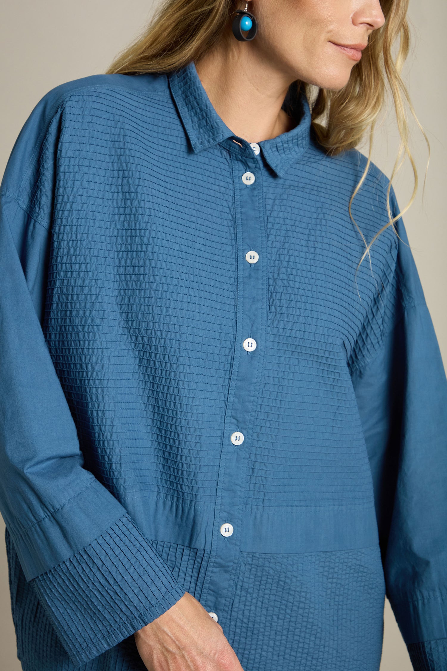 A person wearing a Deep Hem Pleated Shirt in textured blue, adorned with white buttons, and teal earrings embodies a modern classic style as they stand against a neutral background.