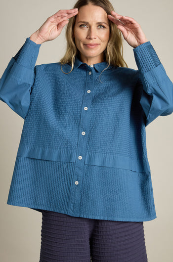 A person stands against a plain background wearing a Yacco Maricard Deep Hem Pleated textured blue shirt and dark pants, raising their hands near their head in a modern classic pose.