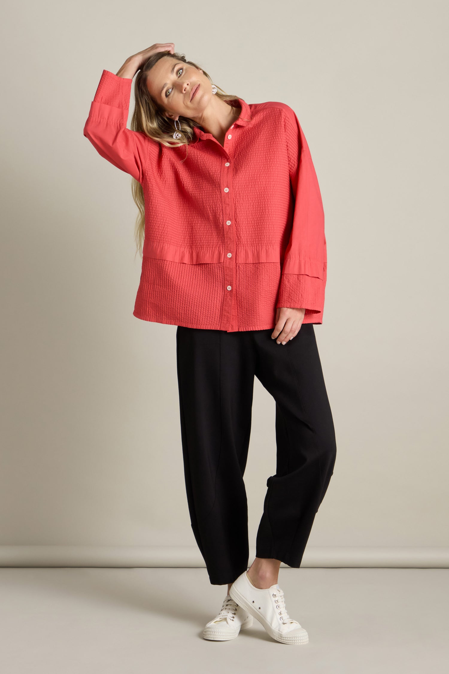 A person stands against a neutral background, wearing a red Deep Hem Pleated Shirt with a boxy fit, paired with black loose-fitting pants and white sneakers, with one hand resting on their head.