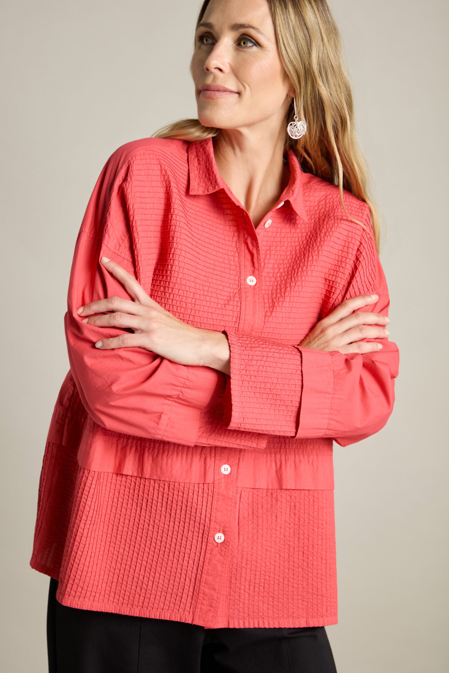 A woman with long blonde hair wears a textured, bright pink Deep Hem Pleated Shirt with a boxy fit by Yacco Maricard. She stands with her arms crossed, looking slightly to the side.