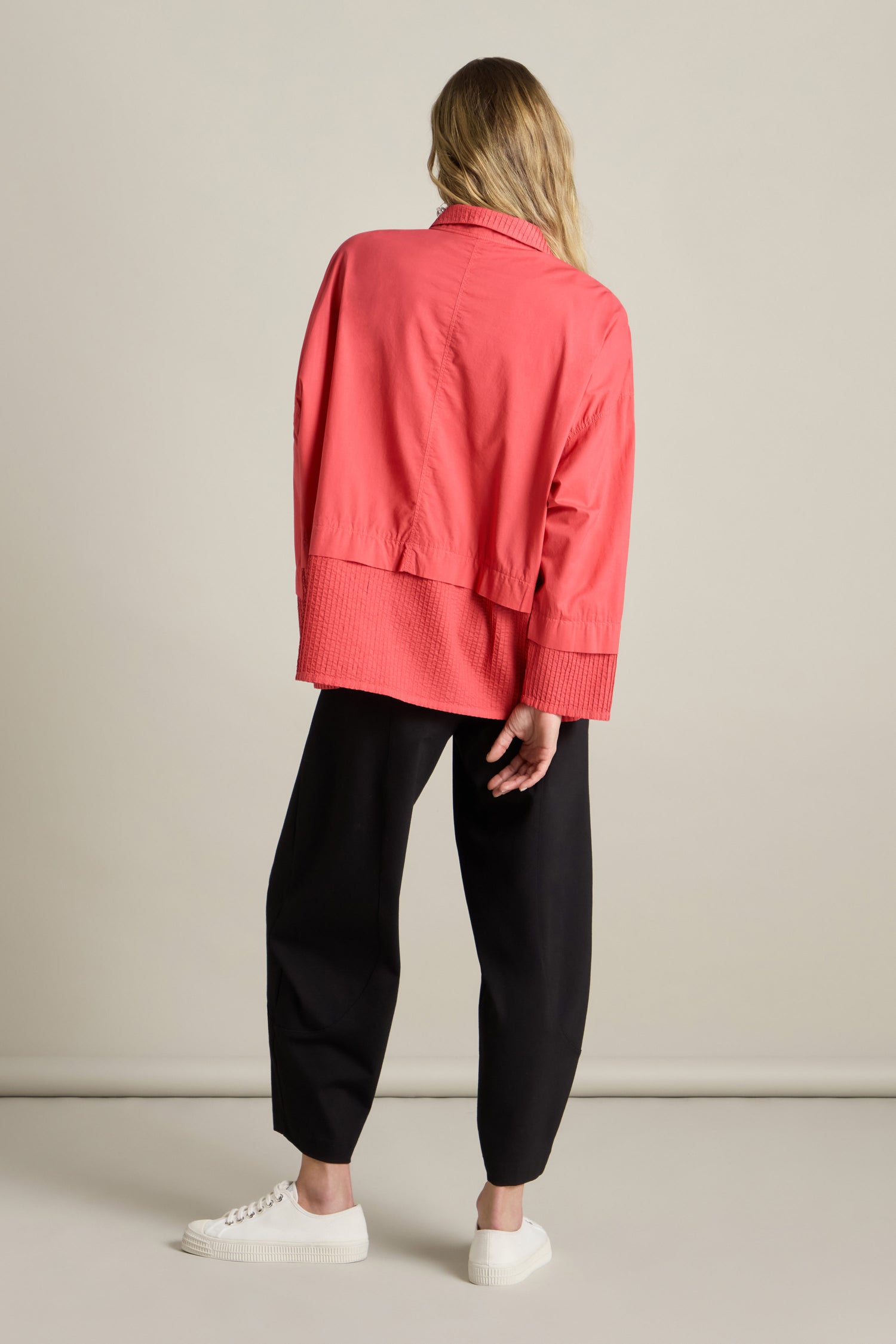 A person with long hair wearing a loose, coral-colored Deep Hem Pleated Shirt by Yacco Maricard with a boxy fit and black pants stands facing away from the camera against a plain background. White sneakers complete the outfit.