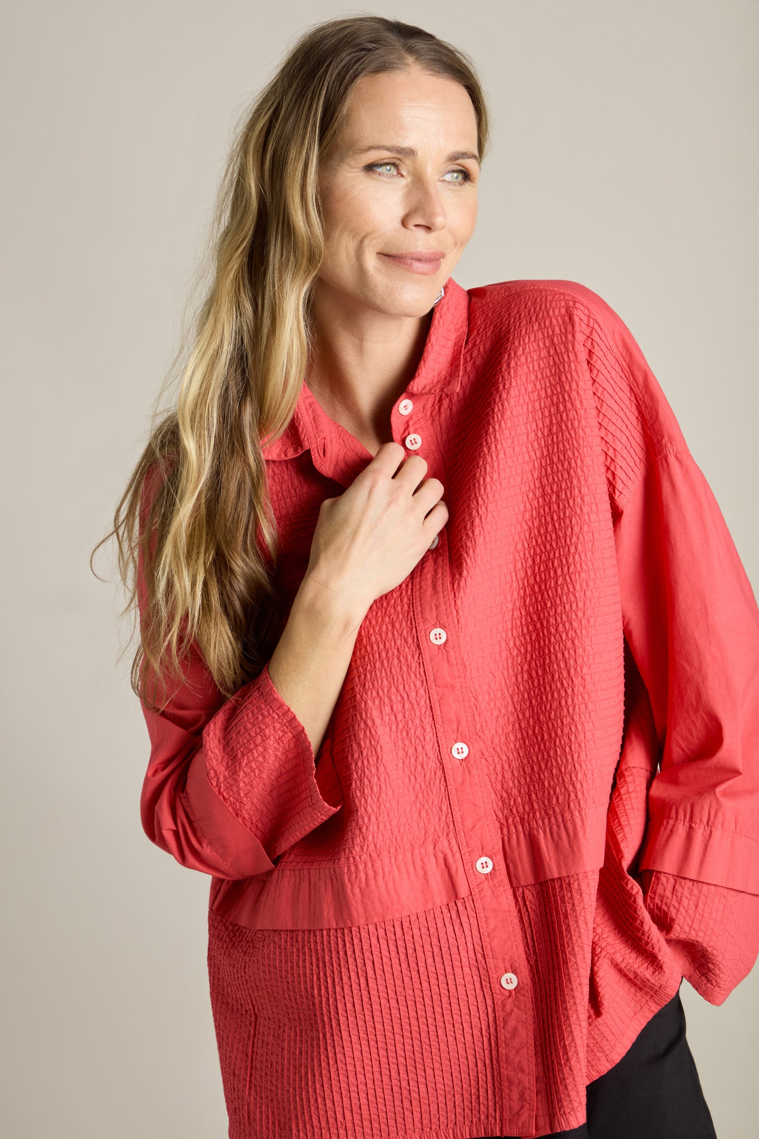 A person with long, blond hair is wearing a red textured Deep Hem Pleated Shirt by Yacco Maricard, smiling slightly while looking off to the side.