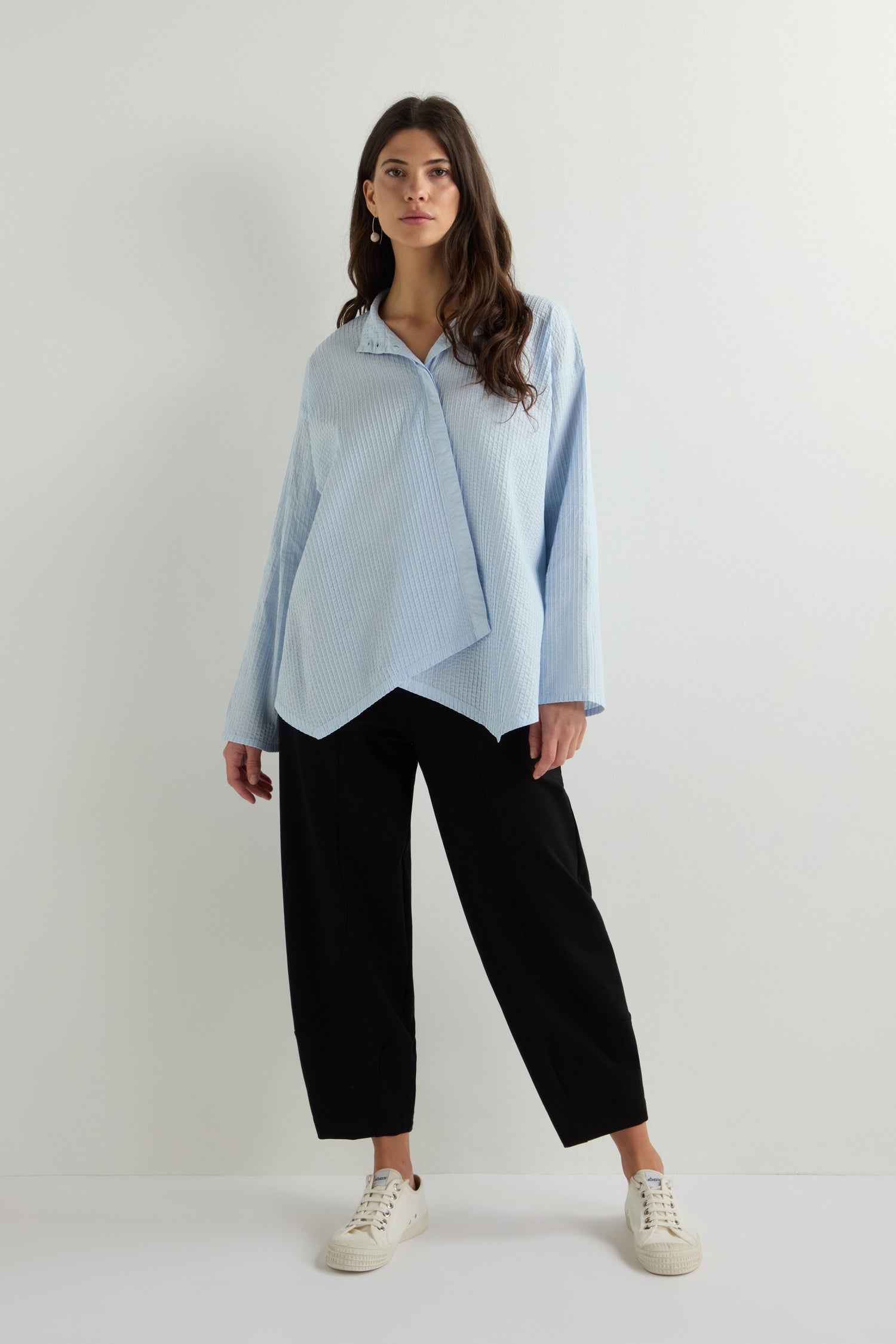 Against a simple backdrop, an individual sports a light blue, oversized Asymmetric Cotton Pleated Shirt from Yacco Maricard with black pants and white sneakers. The airy cotton lawn fabric enhances the outfit's breezy elegance.