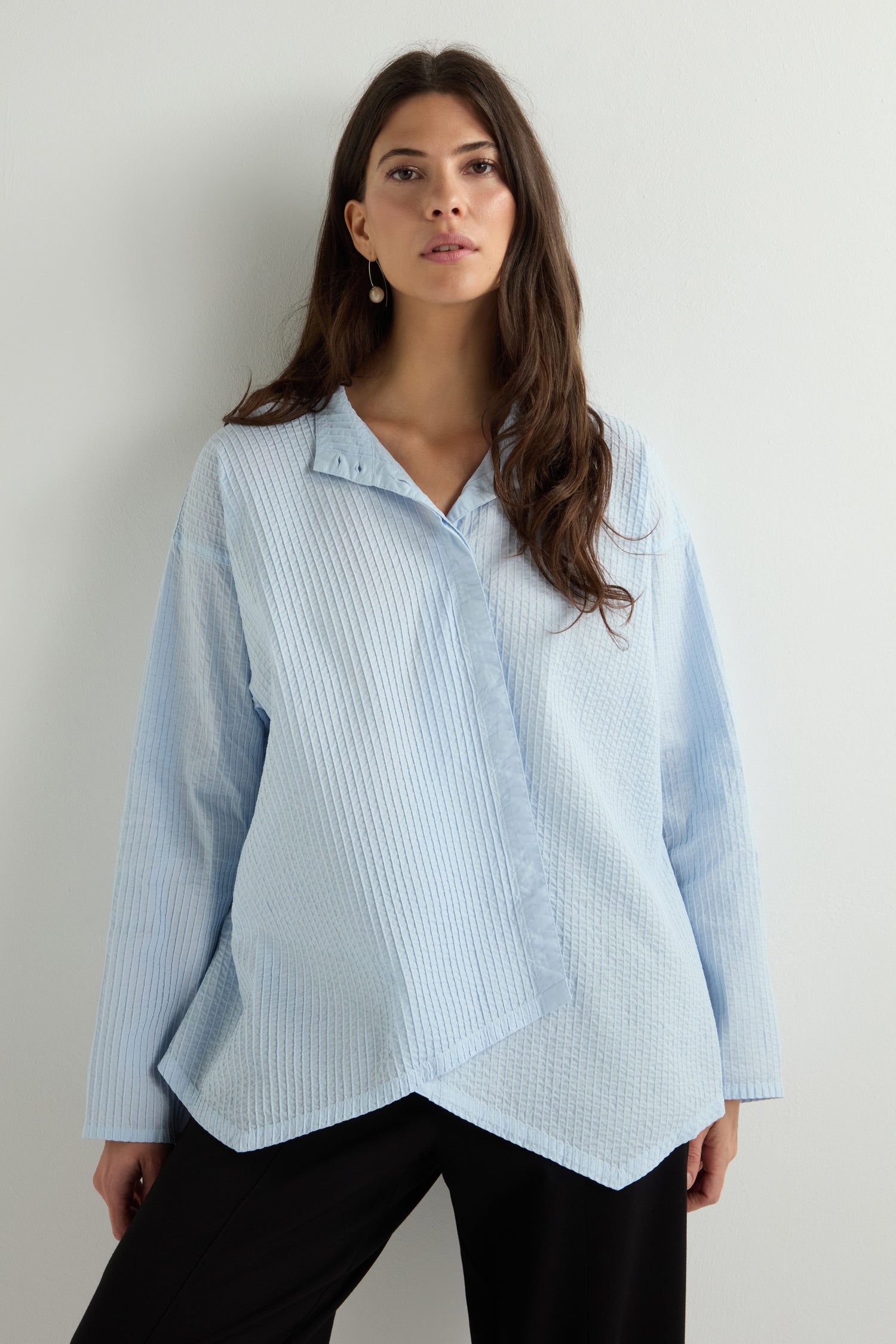 A woman wears a light blue, textured Asymmetric Cotton Pleated Shirt with an asymmetric hem, crafted from cotton lawn, paired with black pants. She stands against a neutral background, echoing the modern elegance of Yacco Maricard's Pleated Short Boxy Shirt design.