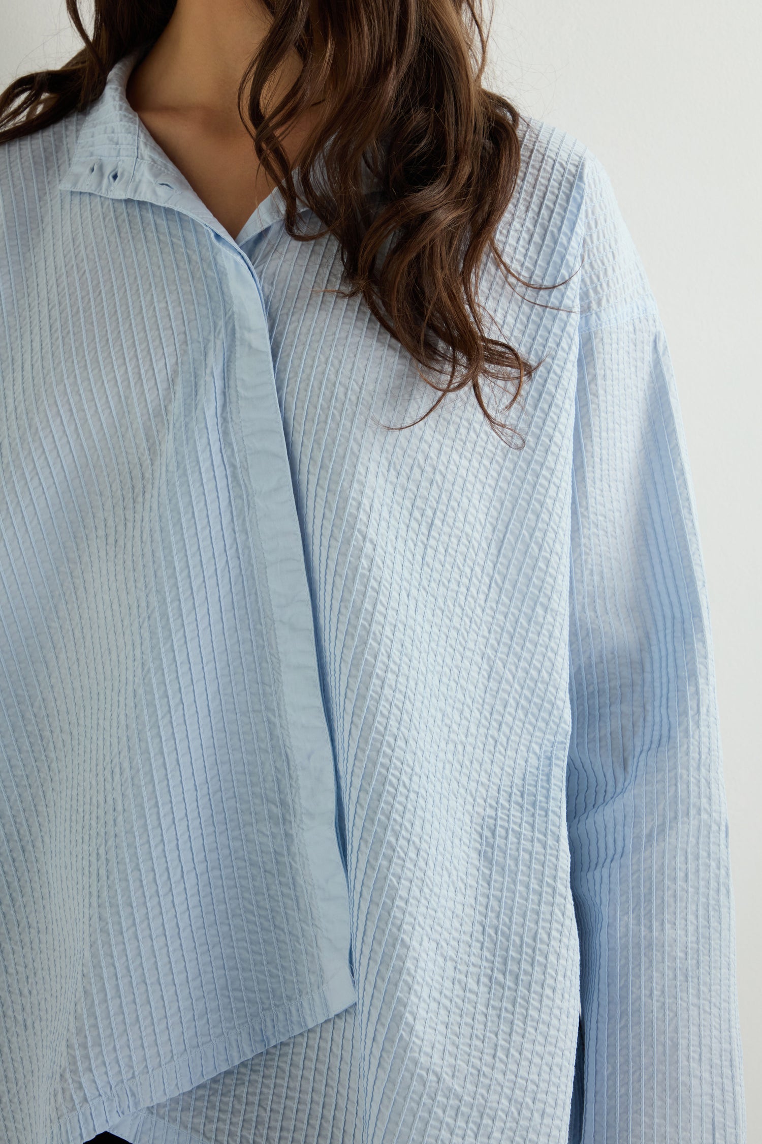 A person with long hair wears an Asymmetric Cotton Pleated Shirt, drawing inspiration from Yacco Maricard, in light blue quilted cotton lawn, standing against a plain white background.