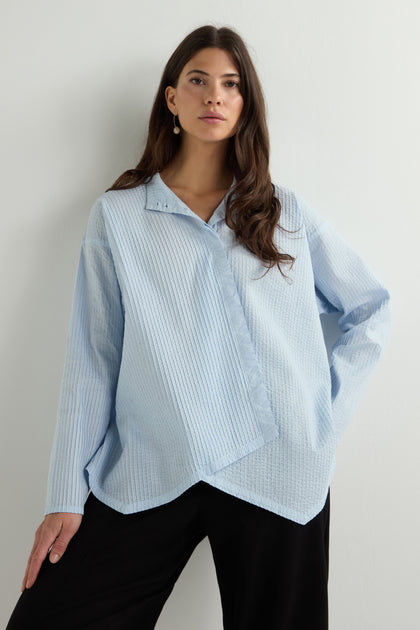 Against a plain backdrop, a person with long hair wears an ensemble reminiscent of Yacco Maricard: an Asymmetric Cotton Pleated Shirt in light blue paired seamlessly with black pants, creating a minimalist yet stylish look.