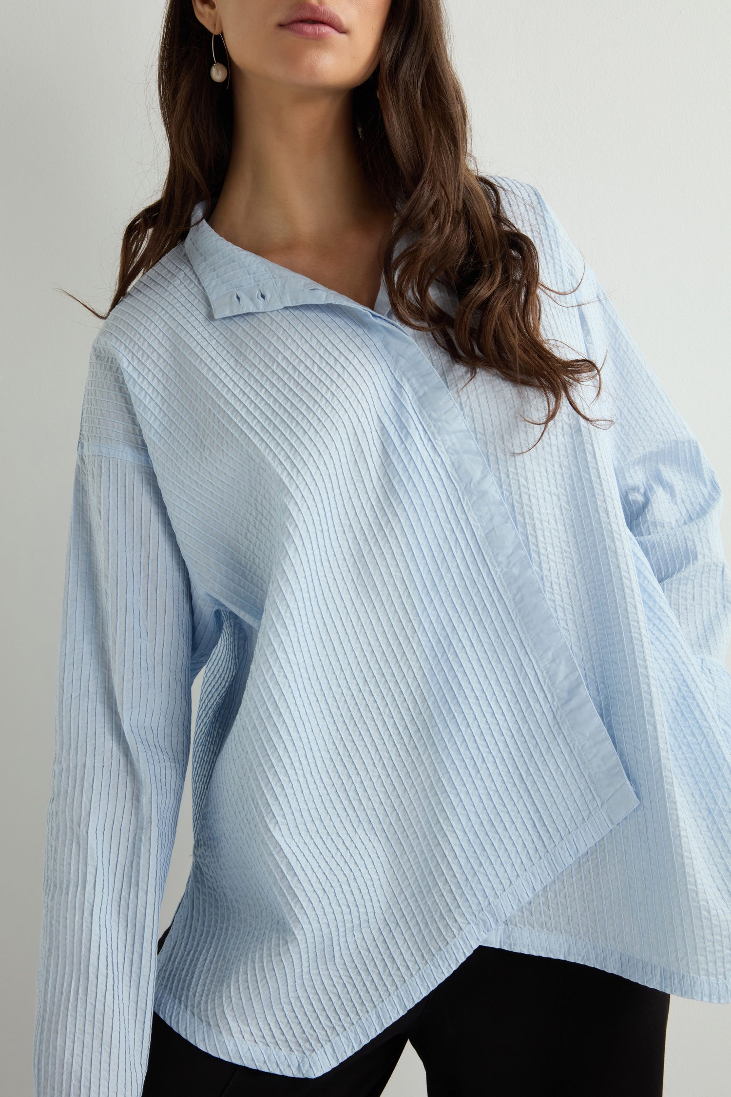 A person is wearing a light blue, textured Asymmetric Cotton Pleated Shirt by Yacco Maricard, made from cotton lawn, paired with black pants against a plain background.