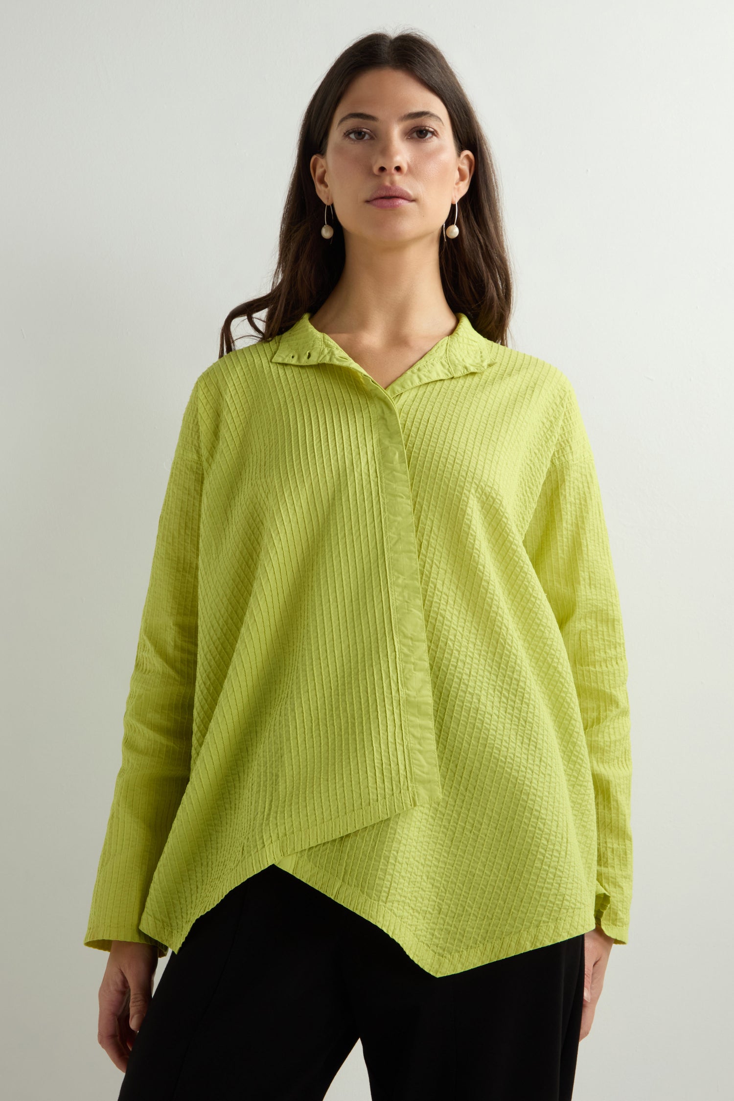 Dressed in a lime green Asymmetric Cotton Pleated Shirt by Yacco Maricard, crafted from cotton lawn, a person pairs it with black pants. They stand confidently against a light background, gazing at the camera.