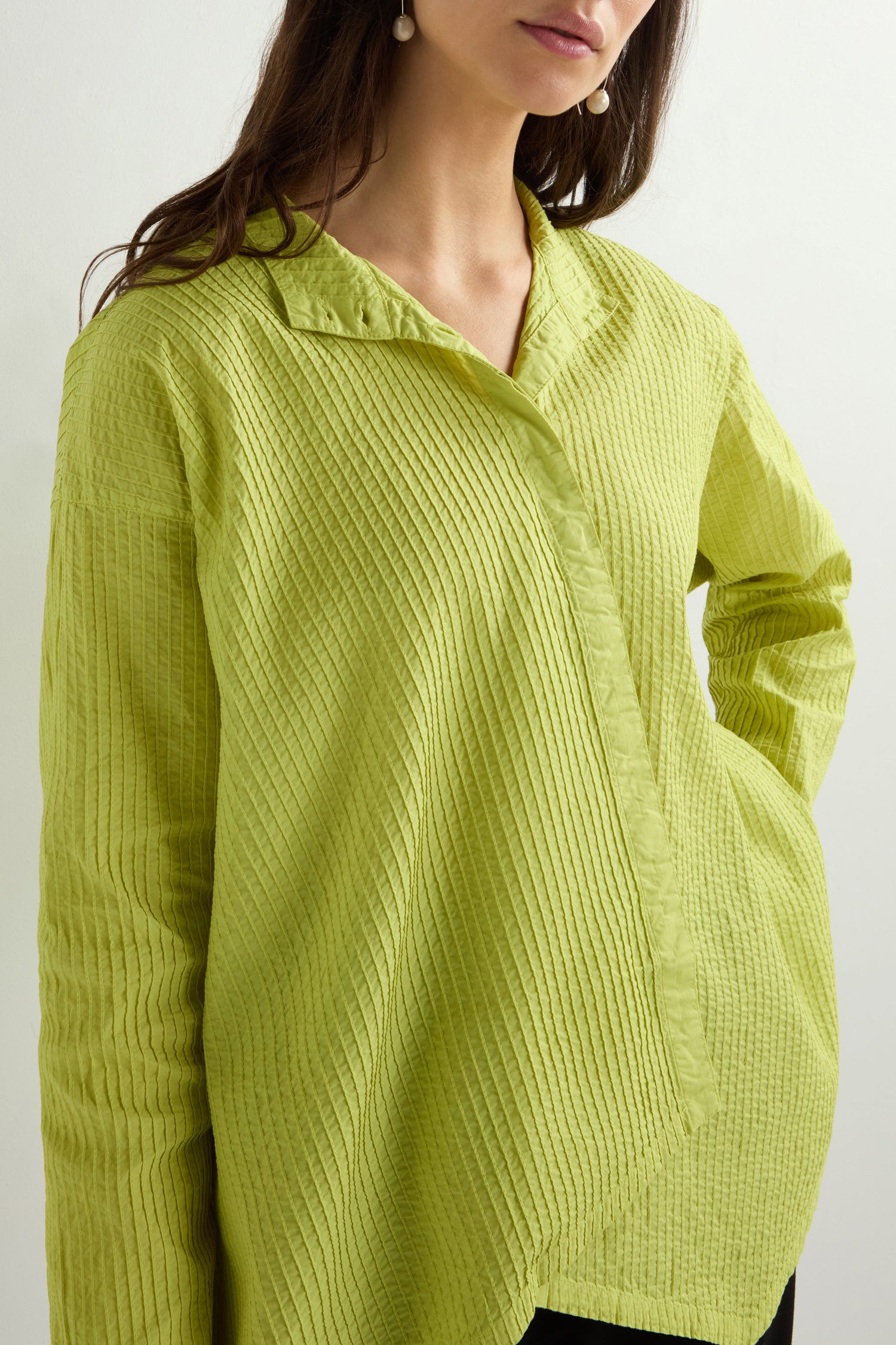 A woman wearing a lime-green Asymmetric Cotton Pleated Shirt by Yacco Maricard, made from cotton lawn, featuring a collar, long sleeves, and a slightly open neckline.