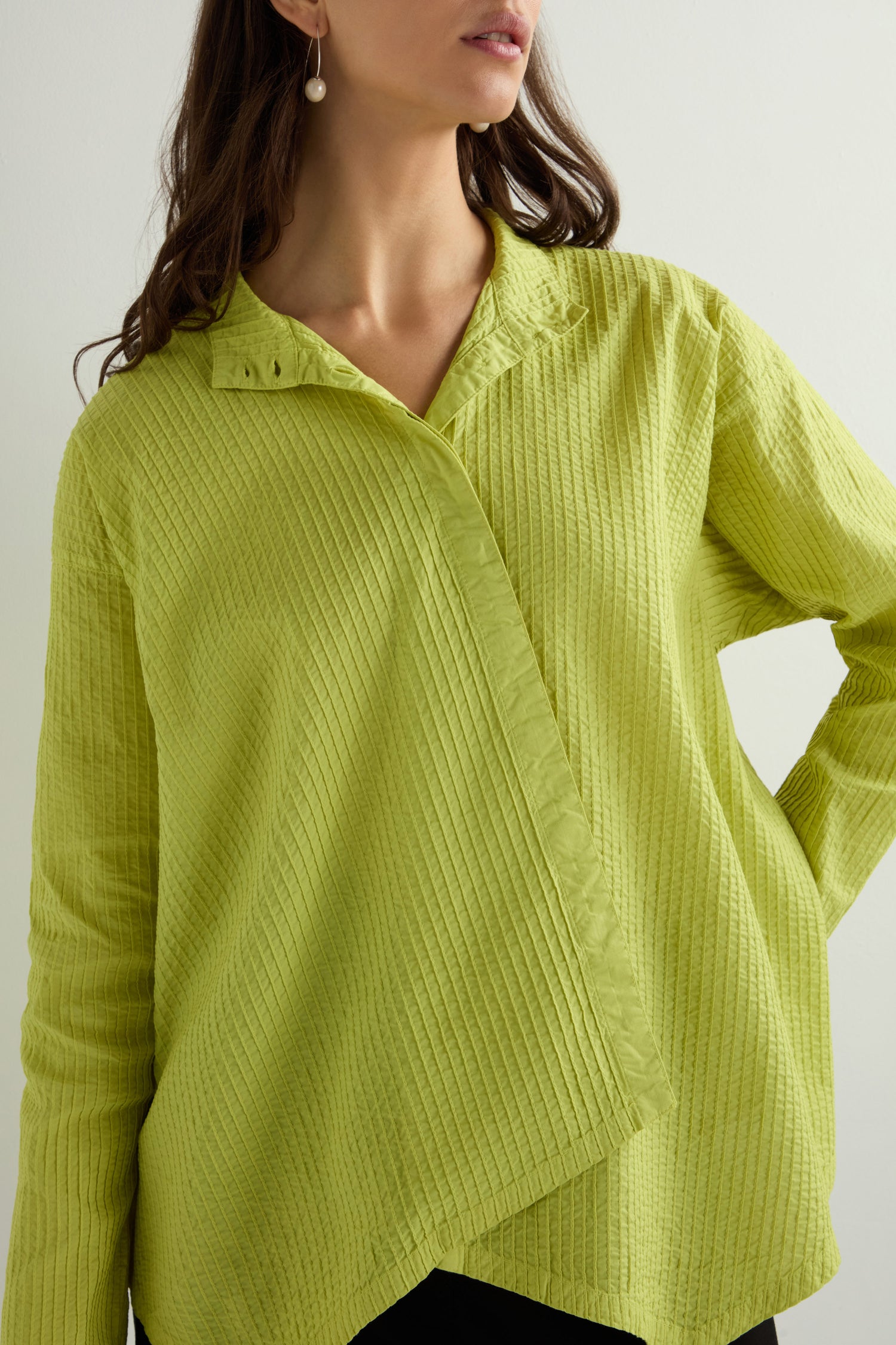 A person in an asymmetric, lime green cotton blouse inspired by Yacco Maricard, paired with pearl earrings, stands against a light backdrop.