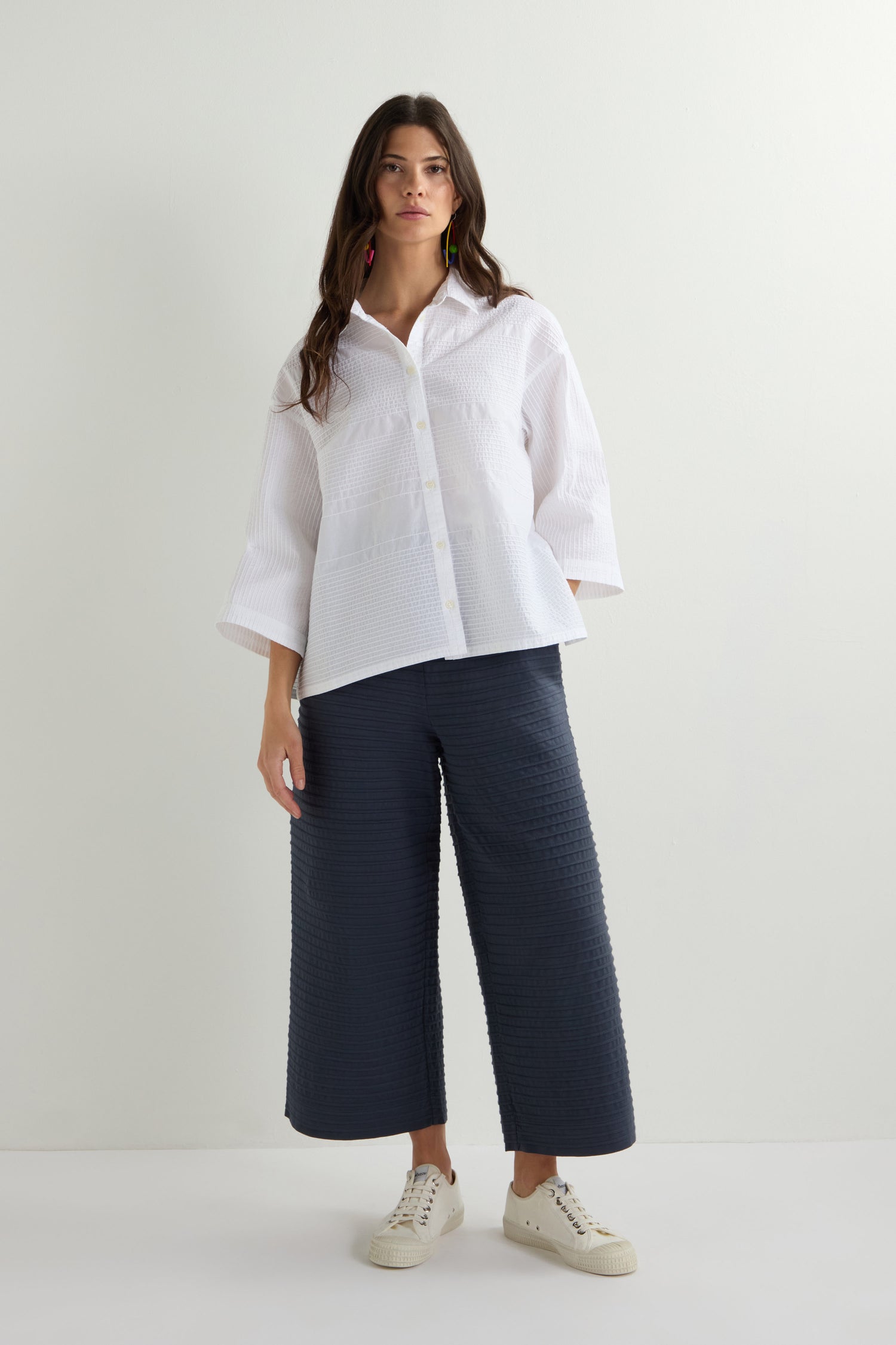 A woman poses against a light background, wearing the Variegated Pleated Shirt—a cotton lawn button-up with pintuck detailing—paired with dark wide-leg pants and white sneakers.