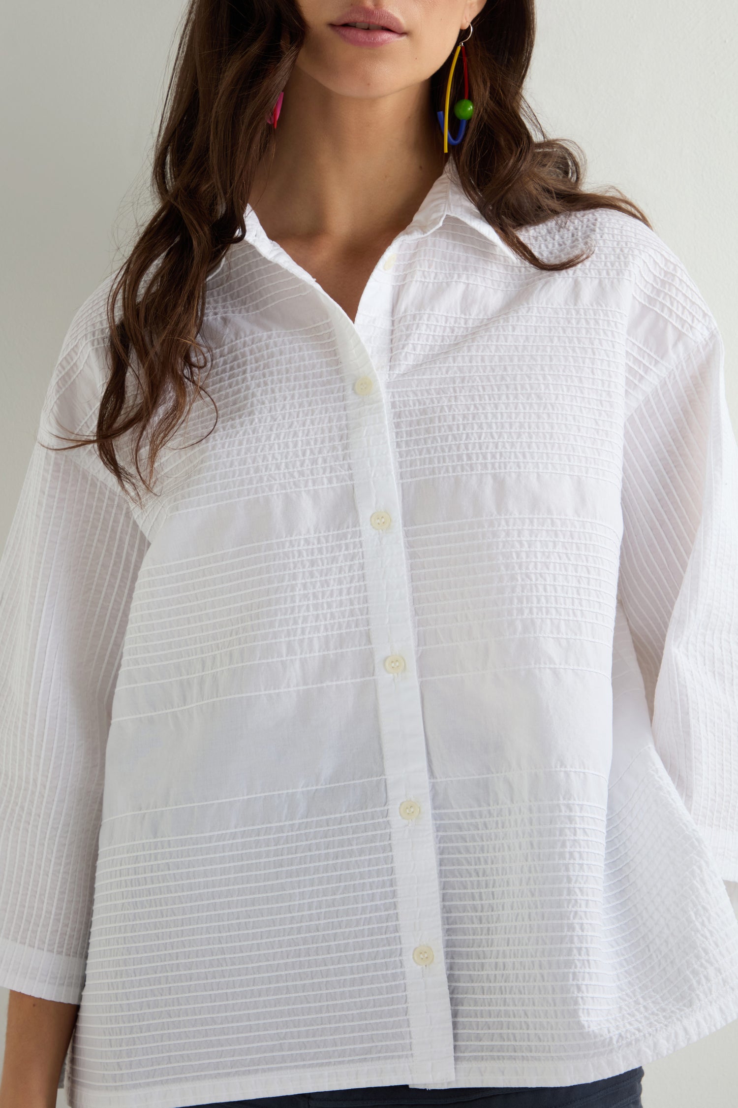 Against a plain backdrop, a person wears the Variegated Pleated Shirt—textured white with pintuck detailing—and vibrant earrings. Their long brown hair cascades down, beautifully contrasting the ensemble's refined simplicity.