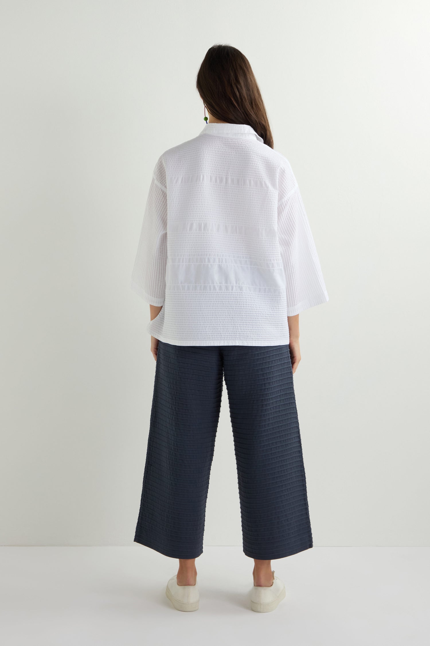 A person stands with their back to the camera, dressed in a white Variegated Pleated Shirt featuring pintuck detailing, paired with dark wide-leg pants and white shoes, set against a plain light background.