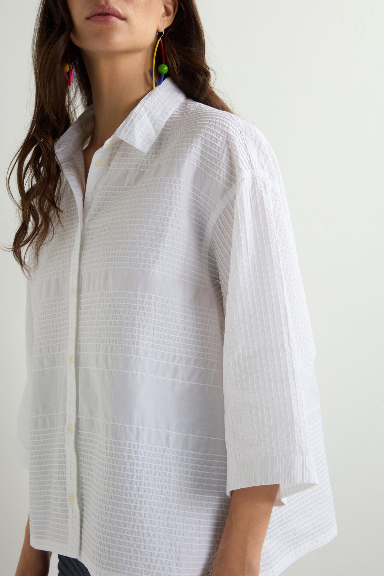 Against a plain background, an individual is dressed in a Variegated Pleated Shirt featuring pintuck detailing and three-quarter sleeves, accompanied by colorful dangling earrings.