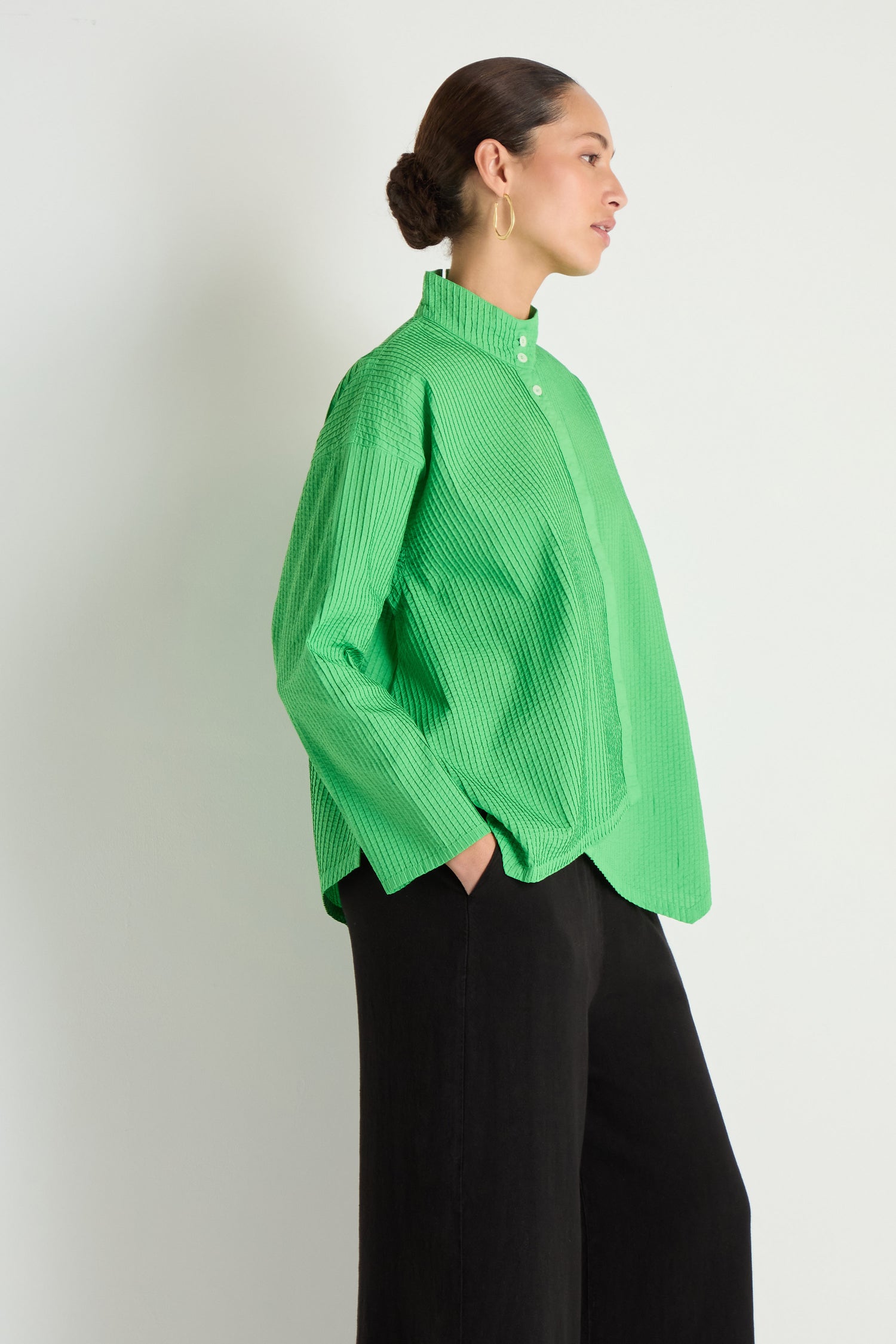 A woman stands in profile wearing the Asymmetric Cotton Bright Green Shirt with a stand neck and textured design, paired with loose black pants, against a plain light background.