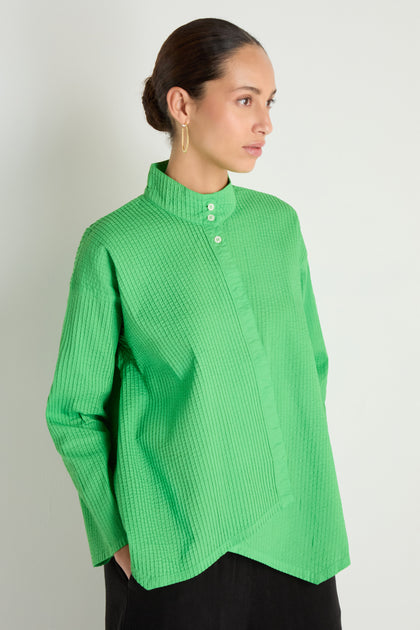Woman in an Asymmetric Cotton Bright Green Shirt featuring a stand collar and white buttons, paired with black pants, standing against a plain light background.