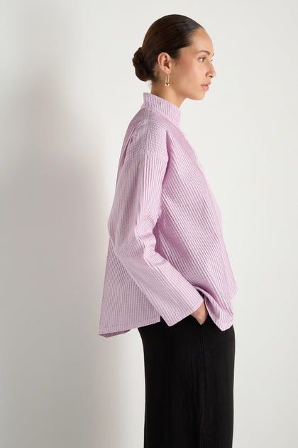 A woman in profile poses against a white backdrop, wearing the Asymmetric Cotton Lilac Shirt with stitched stripes and black pants. The shirt's high collar and her sleek hairstyle add a touch of refined style as she rests one hand in her pocket.