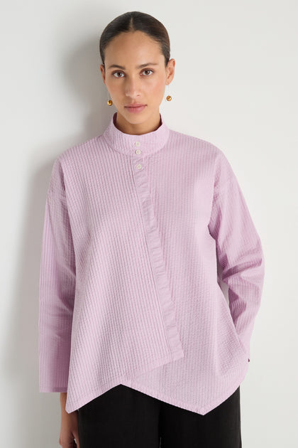 A woman stands against a white wall wearing the Asymmetric Cotton Lilac Shirt, featuring stitched stripes, an asymmetrical hem, buttoned collar, and black pants—an outfit inspired by minimalist Japanese textile design.