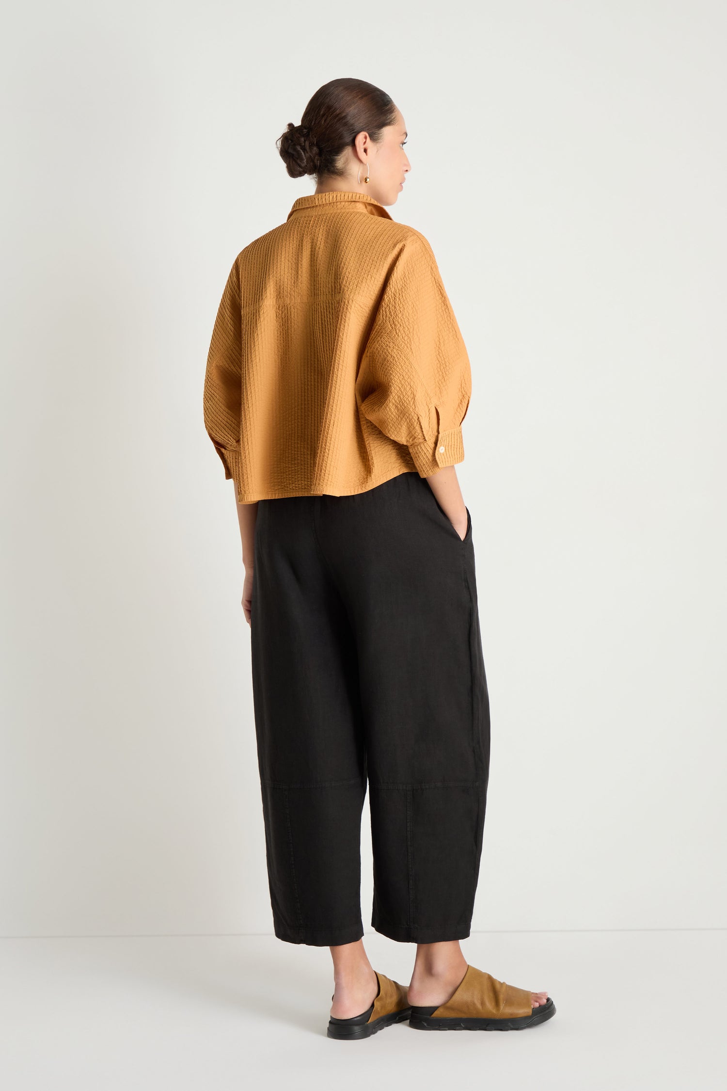 A person stands facing away, wearing the Pleated Sleeve Brown Shirt with a boxy silhouette, paired with black wide-leg pants and tan sandals against a plain white background.