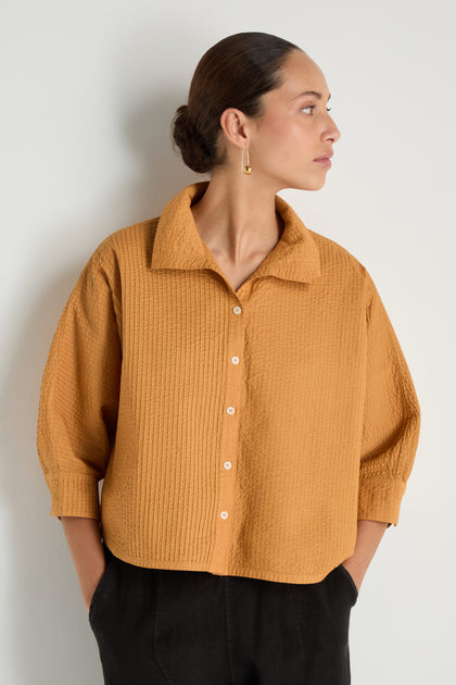 A woman stands against a plain wall, wearing the Pleated Sleeve Brown Shirt with black pants. The shirt’s boxy silhouette and her hands in her pockets create a relaxed, modern vibe as she looks to the side.