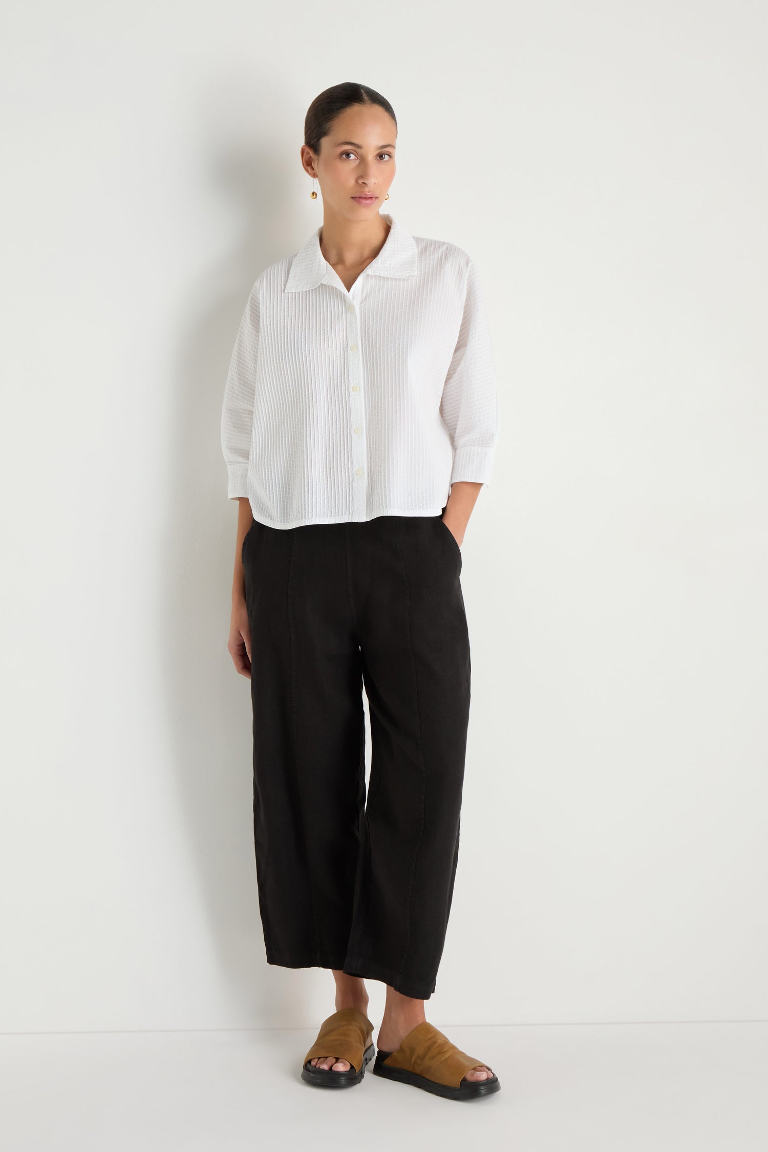 A woman stands against a white wall wearing the Pleated Sleeve White Shirt, styled with black cropped pants and brown sandals.
