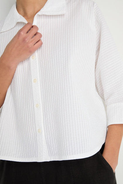 Person wears the Pleated Sleeve White Shirt, a textured cotton button-up with three-quarter pleated sleeves and a boxy silhouette. One hand is near the collar; the other rests in a pocket.