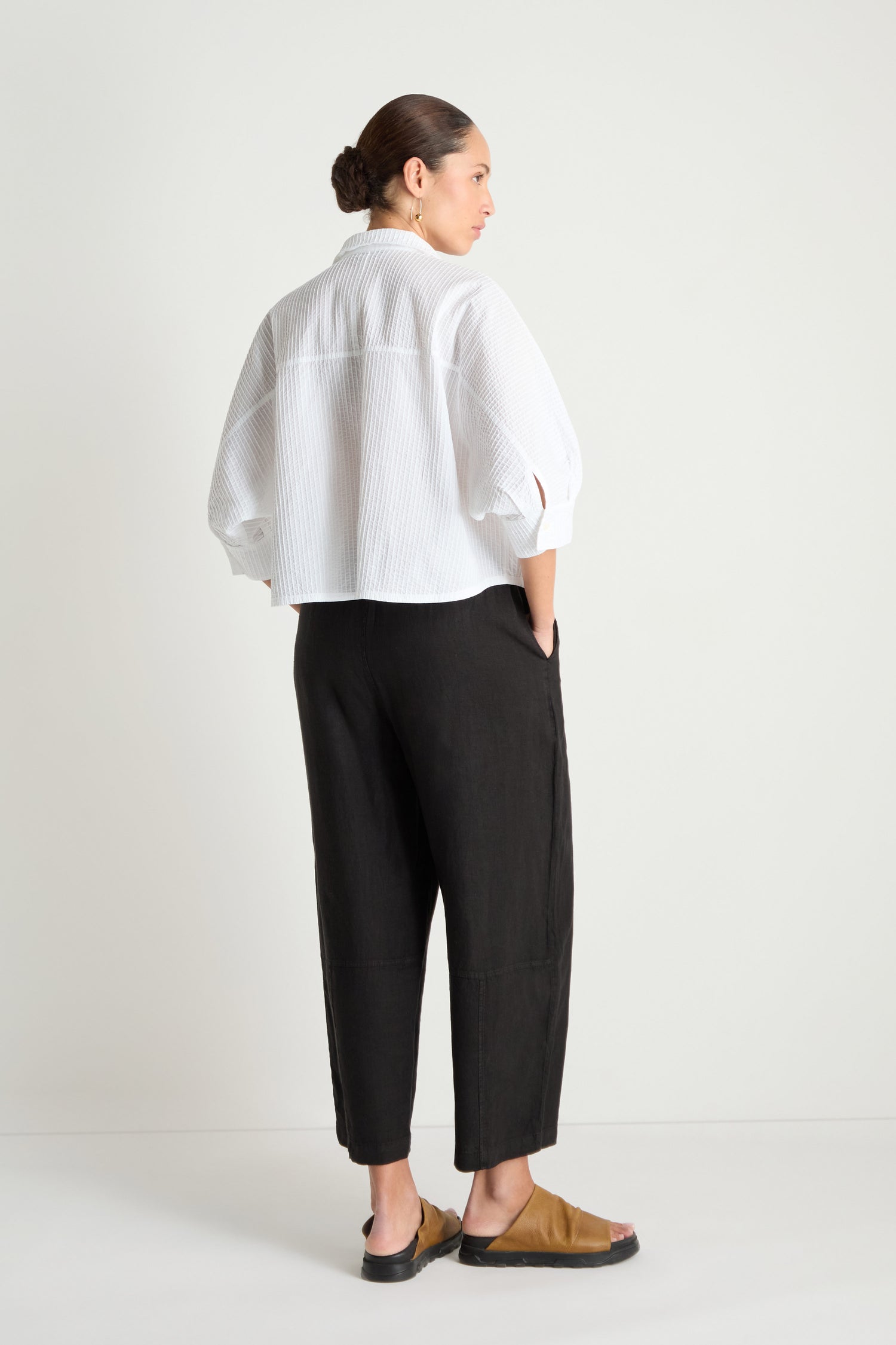 A person stands with their back to the camera, wearing the Pleated Sleeve White Shirt, black loose trousers, and tan slide sandals against a plain light background.