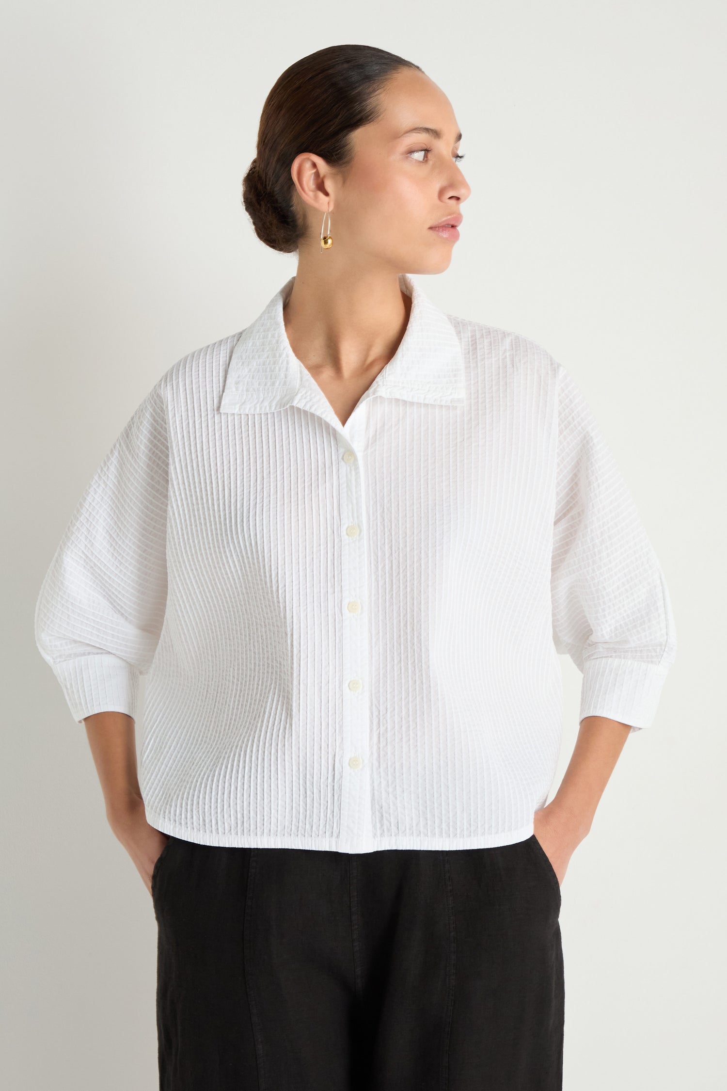 Wearing the Pleated Sleeve White Shirt paired with black pants, a person stands against a plain background, hands in pockets and looking to the side, highlighting its modern boxy silhouette.