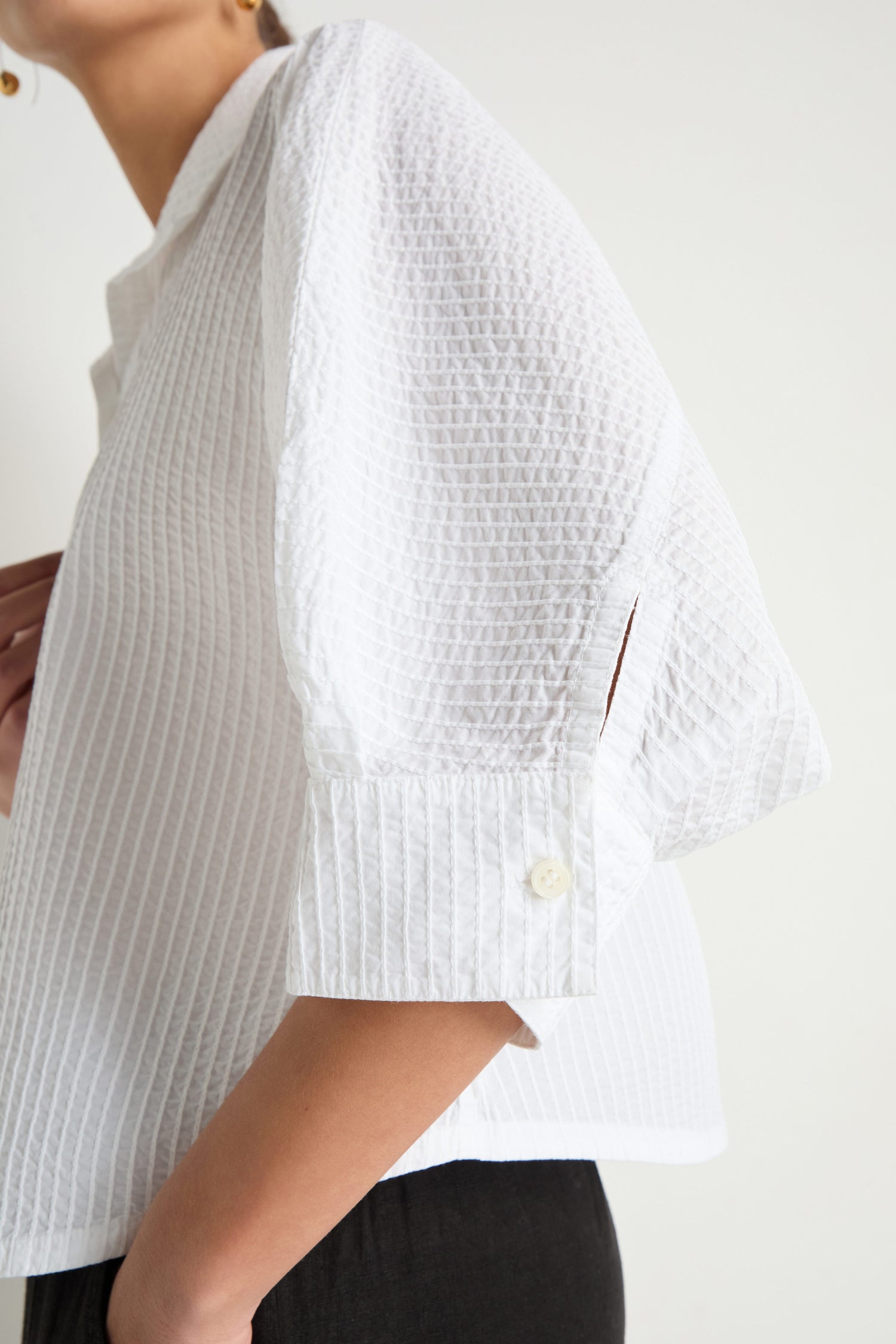 A person shown from shoulders to waist wears the Pleated Sleeve White Shirt, featuring textured white cotton and three-quarter sleeves, paired with black pants.