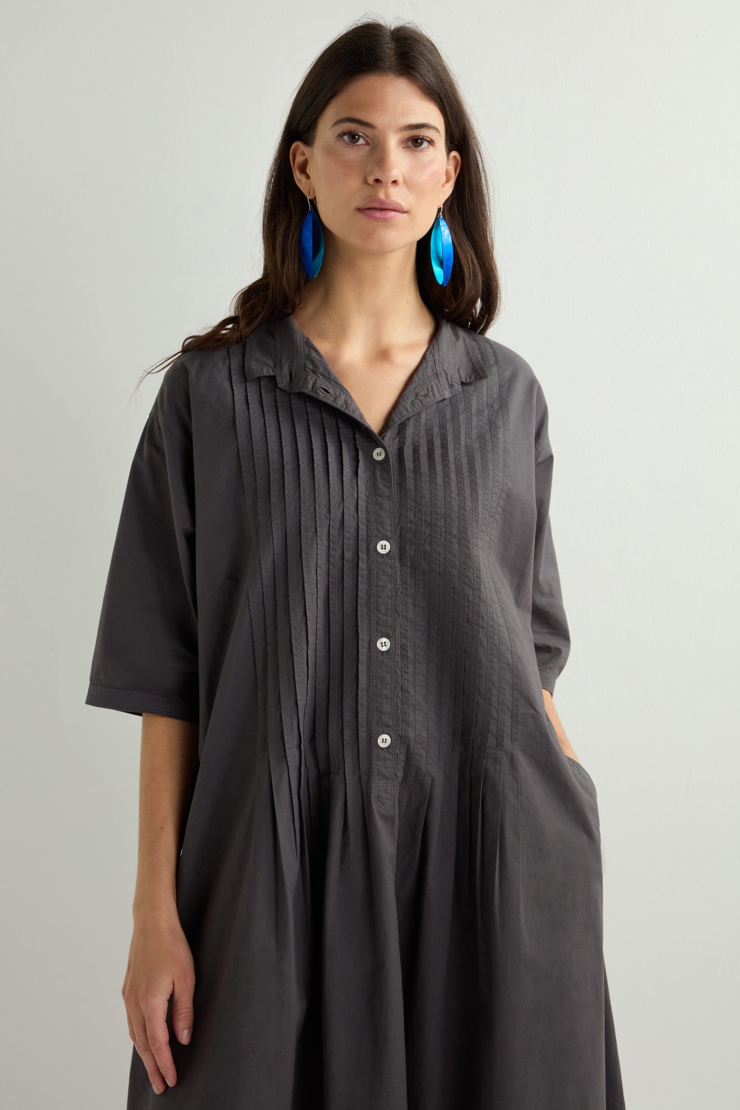 A person stands against a plain background in a gray Cotton Pleat Detail Dress with vertical pleats and a relaxed, oversized fit, paired with striking blue earrings.