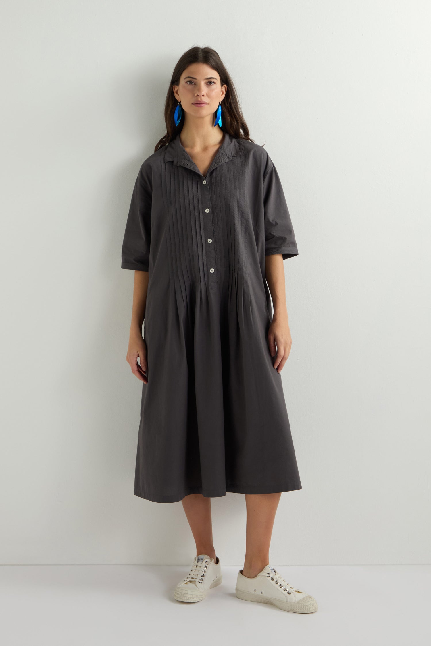 A person poses against a white background wearing a Cotton Pleat Detail Dress in dark grey with an oversized fit, complemented by blue earrings and white sneakers.