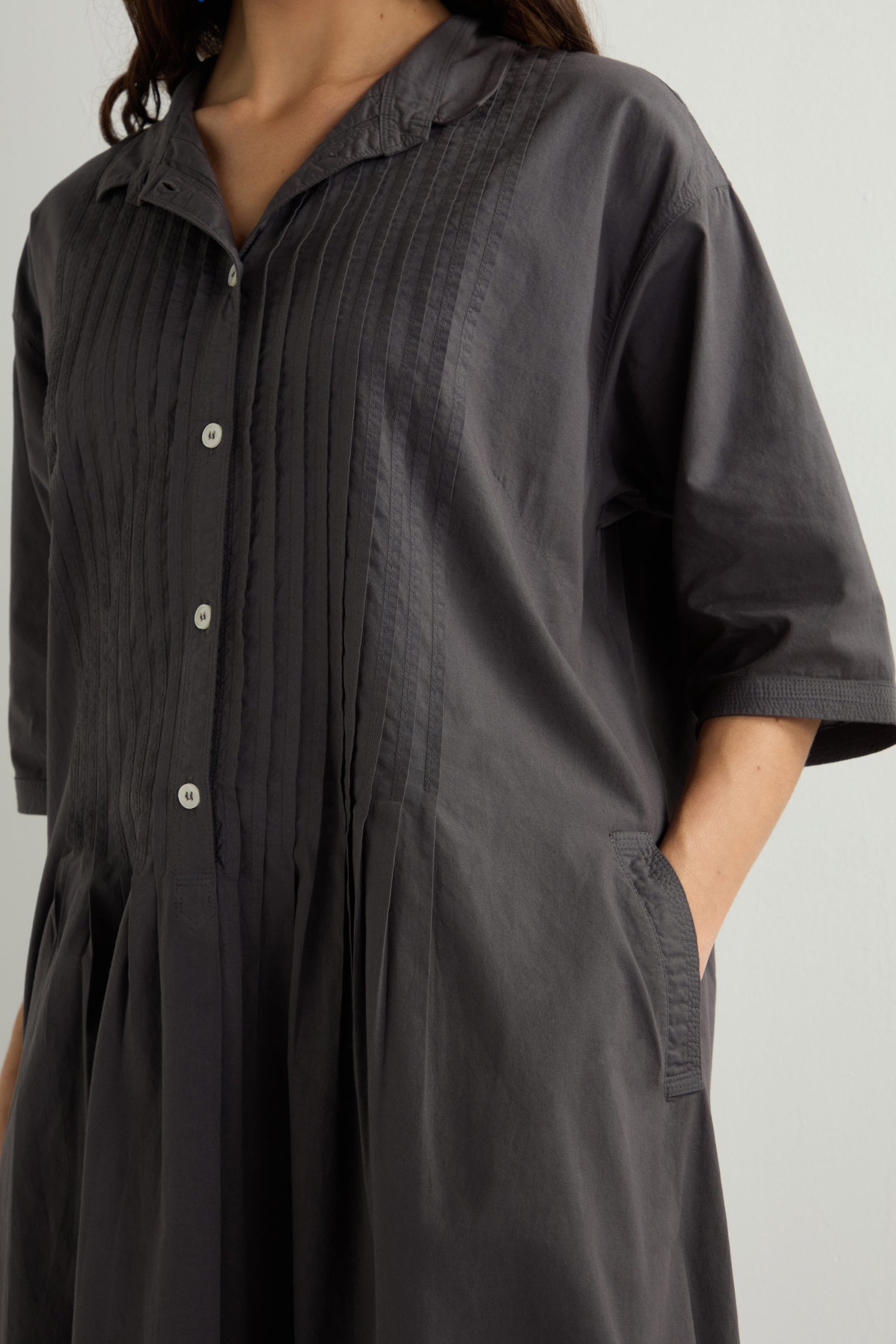 Someone stands against a plain background wearing a dark gray Cotton Pleat Detail Dress, which has short sleeves and a relaxed, oversized fit.