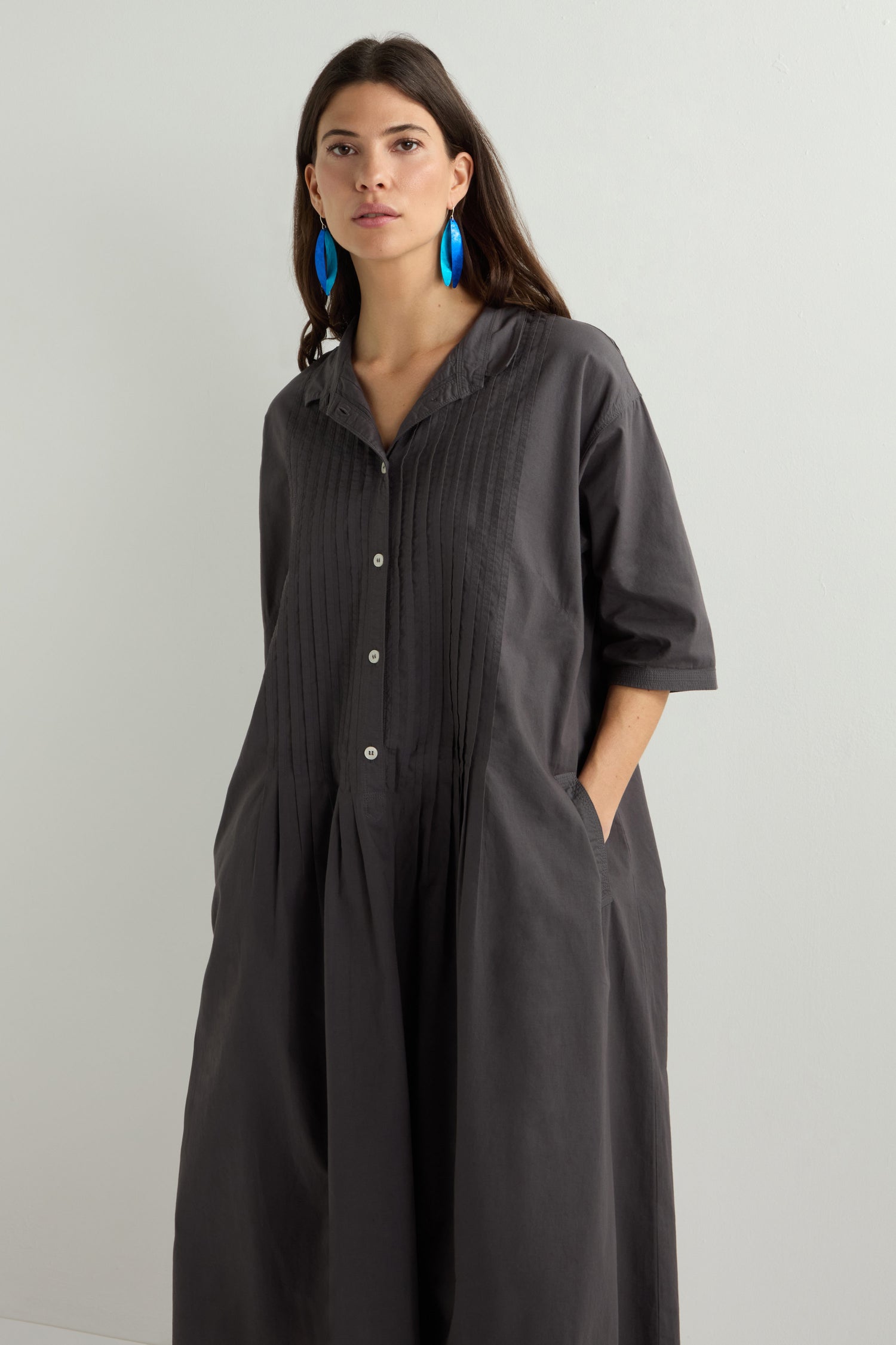 A woman in a gray Cotton Pleat Detail Dress, known for its relaxed oversized fit, stands with her hands in pockets against a light background, pairing it stylishly with large blue earrings.