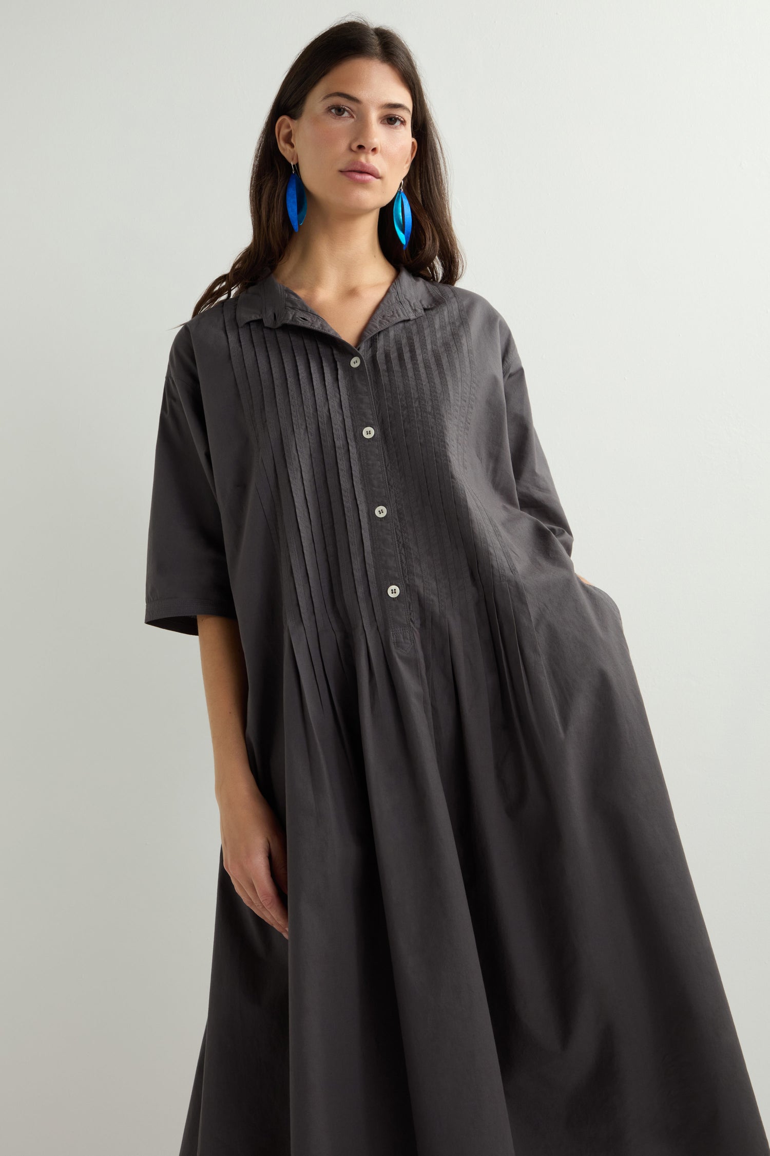 The woman wears a Cotton Pleat Detail Dress, featuring a relaxed oversize fit with unique garment-dye pleats and buttons. Her long dark hair and large blue earrings stand out against a plain light background.