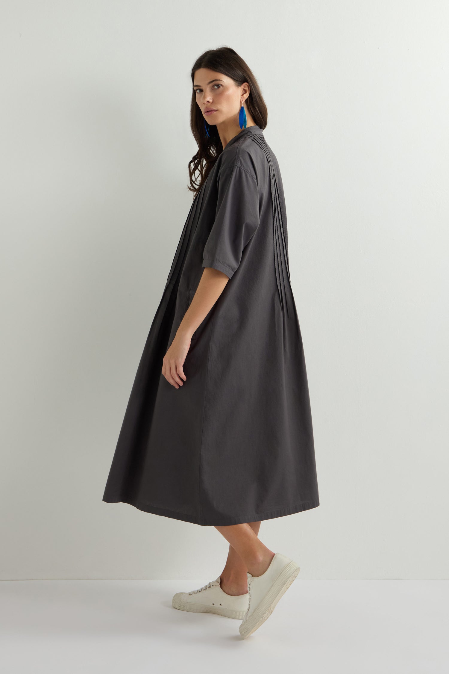 A person wearing a gray Cotton Pleat Detail Dress, paired with blue earrings and white sneakers, stands against an off-white background, looking to their left.