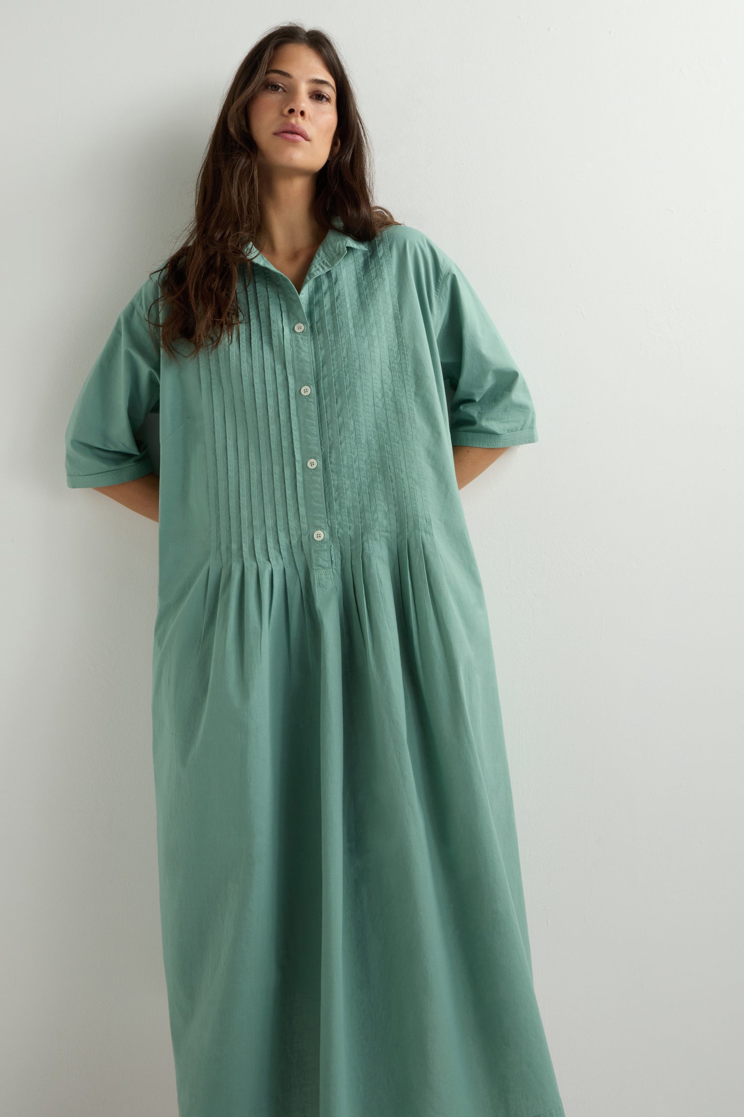 A woman poses against a white background, wearing an oversized green Cotton Pleat Detail Dress, enhanced by a garment-dye finish.