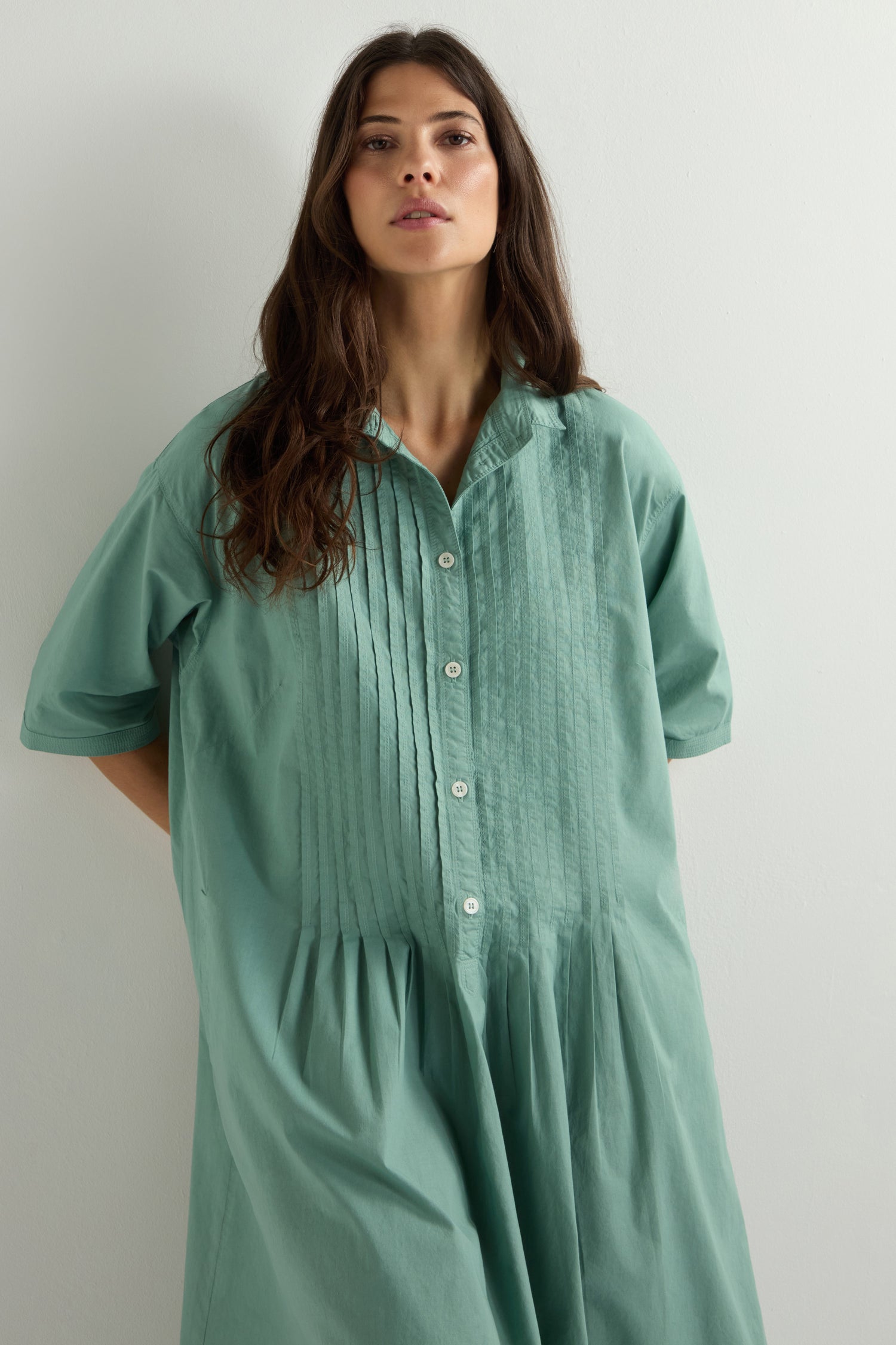 Against a white wall, a person with long dark hair and a neutral expression wears an oversized Cotton Pleat Detail Dress in light green, crafted using the garment-dye process.