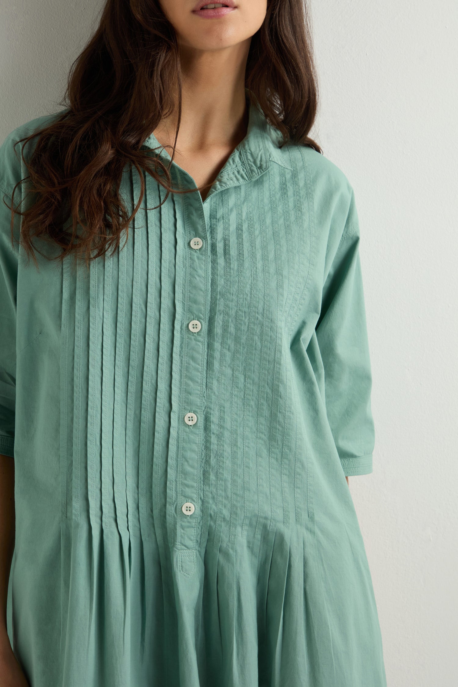 A woman in a teal Cotton Pleat Detail Dress, crafted through garment-dyeing, stands against a white background.