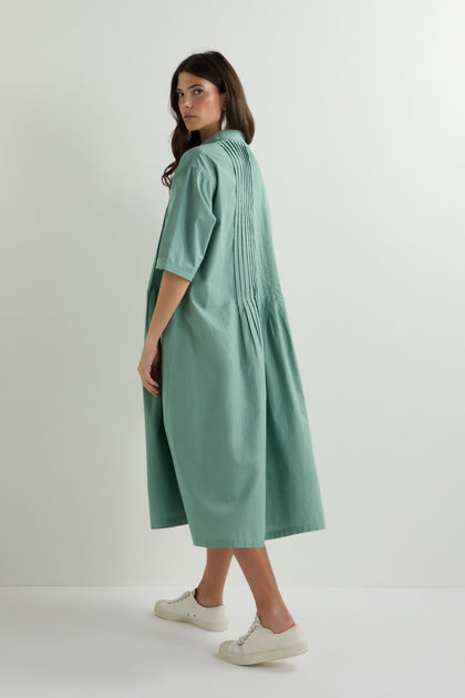 A woman with long hair stands sideways in a knee-length, teal Cotton Pleat Detail Dress, featuring an oversized fit and garment-dye process. She pairs it effortlessly with white sneakers against a plain white background.