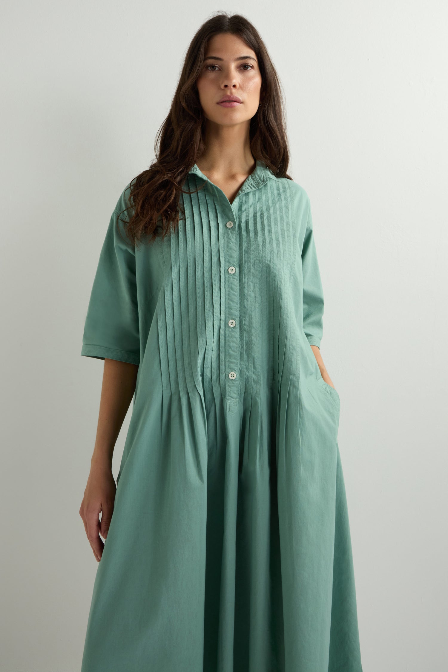 A woman in a green Cotton Pleat Detail Dress, known for its relaxed oversized fit, stands against a plain white backdrop.