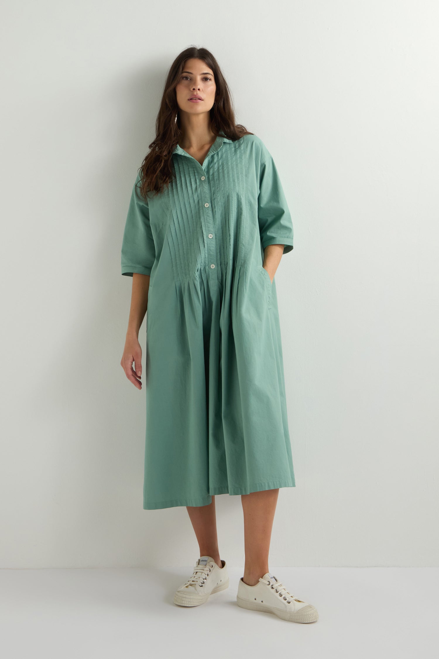 A woman wearing a green Cotton Pleat Detail Dress, featuring an oversized fit and pockets, stands against a plain white background paired with white sneakers.