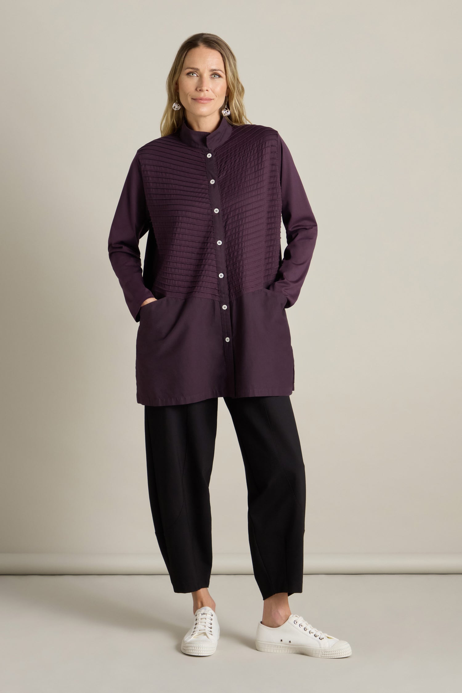 The person stands against a plain background, wearing the Cotton Jersey And Calico Overshirt in dark purple with an embroidered placket, black pants, and white sneakers, with hands in pockets.